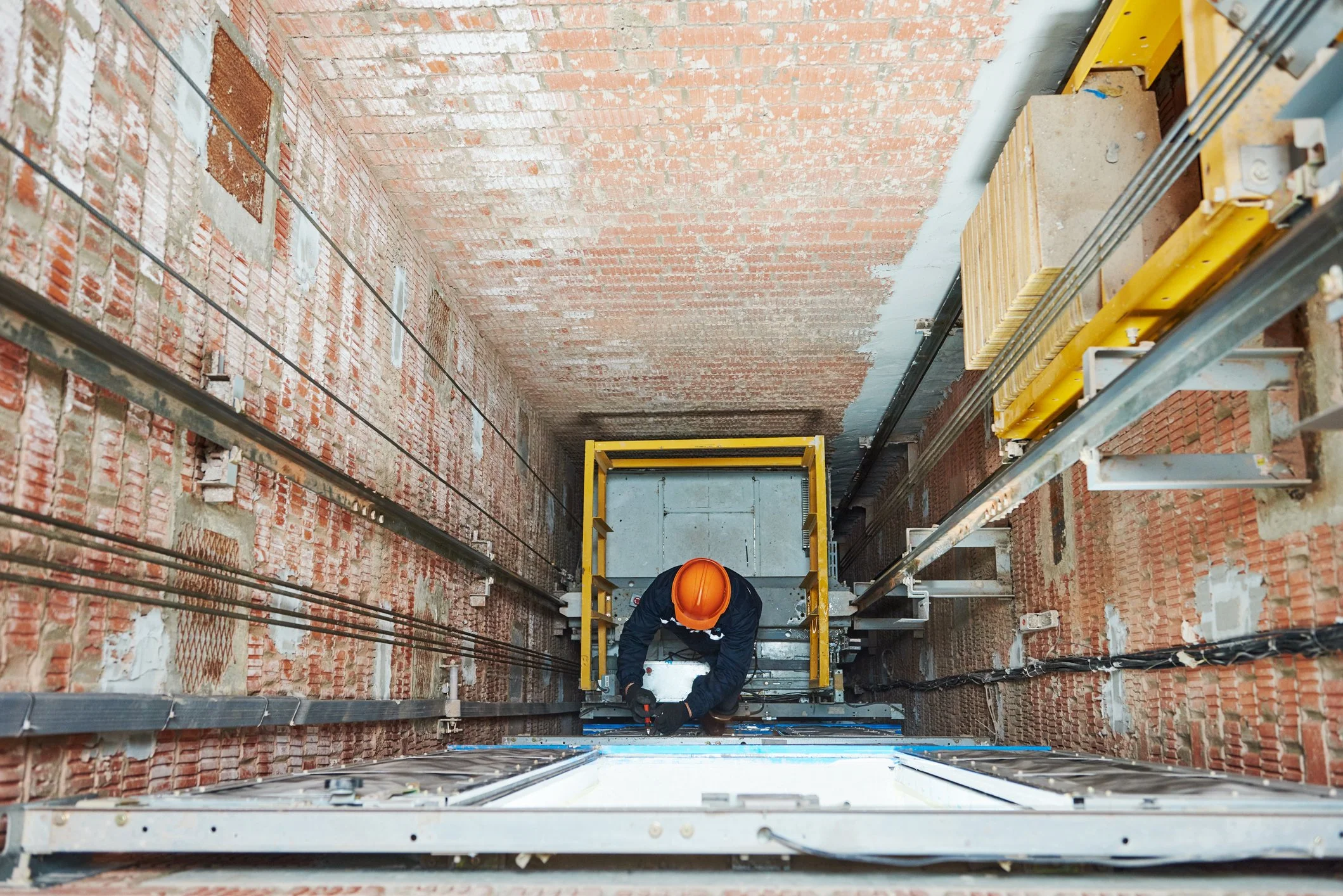 View from the top of a construction lift showing brick walls with exposed wires and an overhead worker wearing an orange safety helmet working on the ceiling.