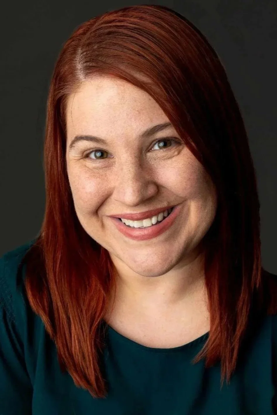 Close-up of a smiling woman with red hair and green eyes against a dark background.