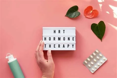 Hand holding a lightbox with the words 'HRT HORMONE THERAPY,' surrounded by hearts, a blister pack of pills, and a small green bottle on a pink background.
