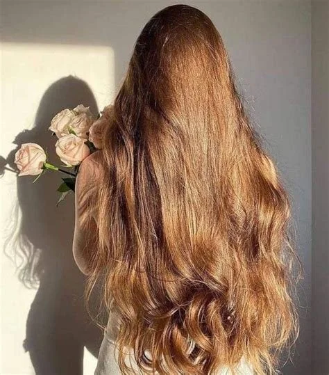 A woman with long, wavy, reddish-brown hair holding a bouquet of pink roses, with her back facing the camera, and sunlight casting a shadow on the wall.