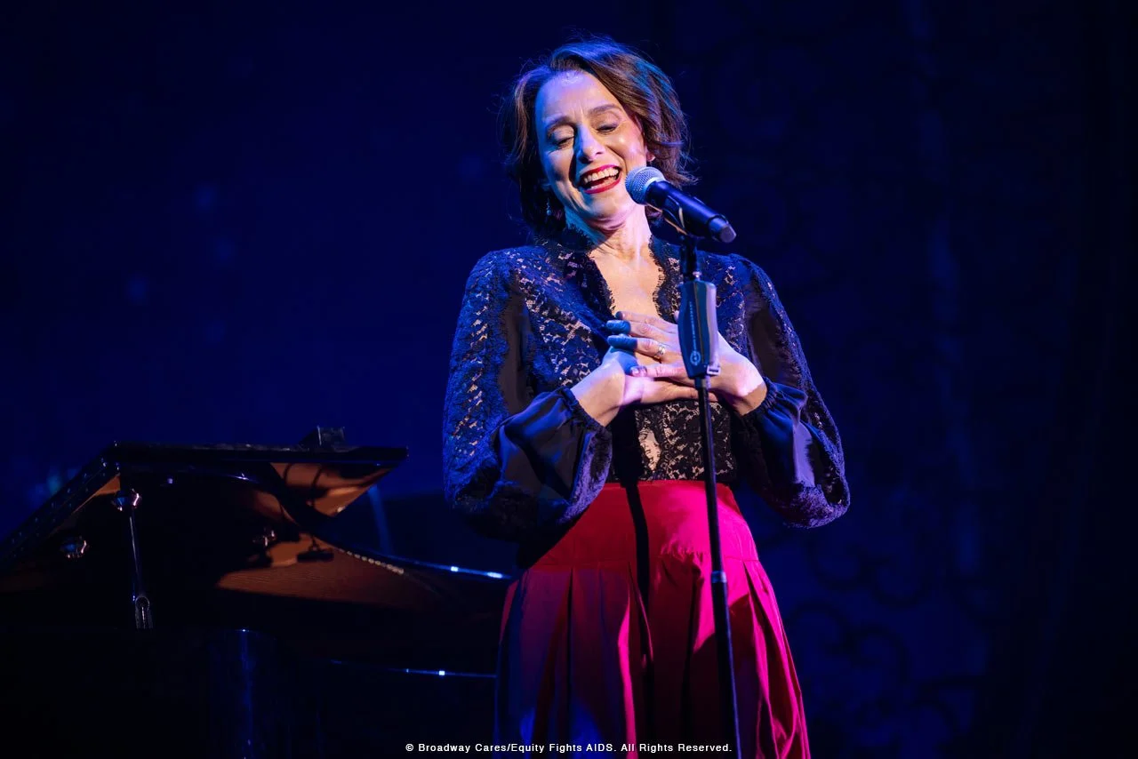 A woman singing into a microphone on stage, wearing a black lace top and red skirt, with a grand piano in the background.