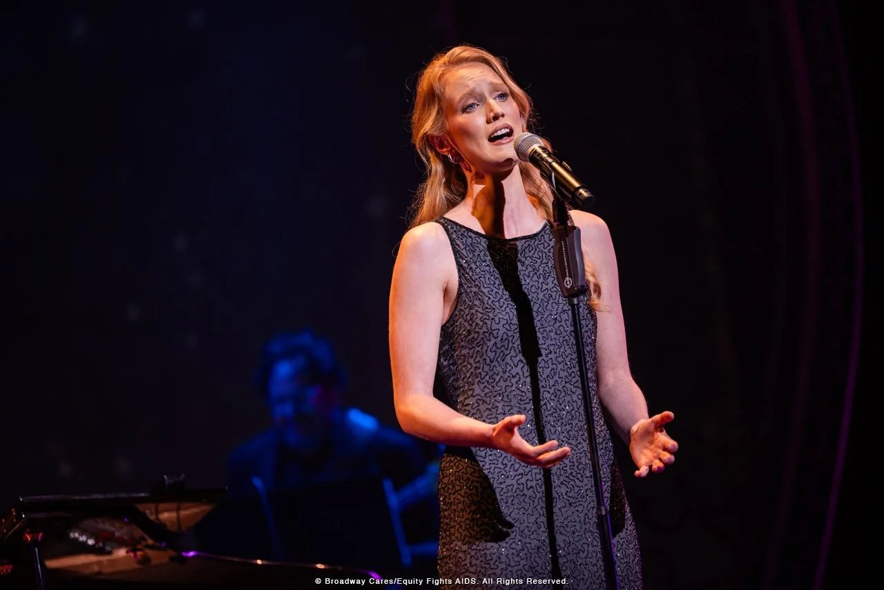 A woman singing passionately into a microphone on a stage, with a pianist in the background.