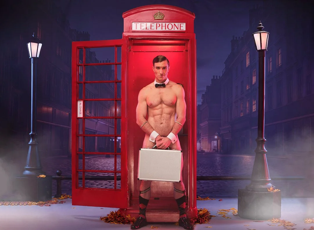 A shirtless man wearing a bow tie, cuffs, and socks with a briefcase stands in a red British telephone booth on a foggy city street at night.