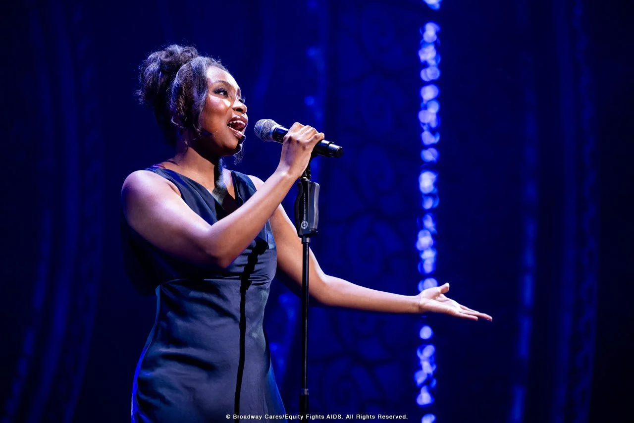 A woman singing into a microphone on stage, with blue curtains in the background.