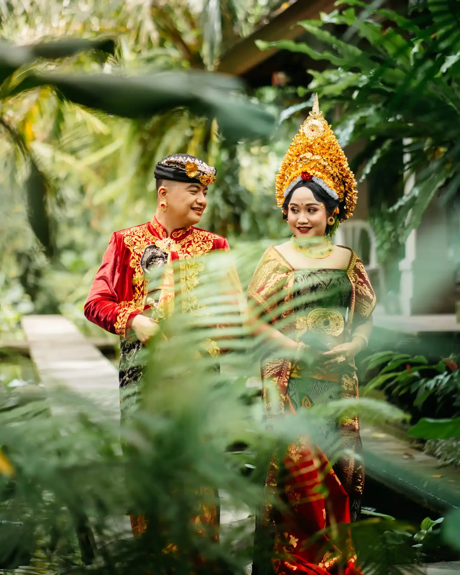 A couple dressed in traditional Indonesian wedding attire, standing amidst lush greenery and smiling at each other.