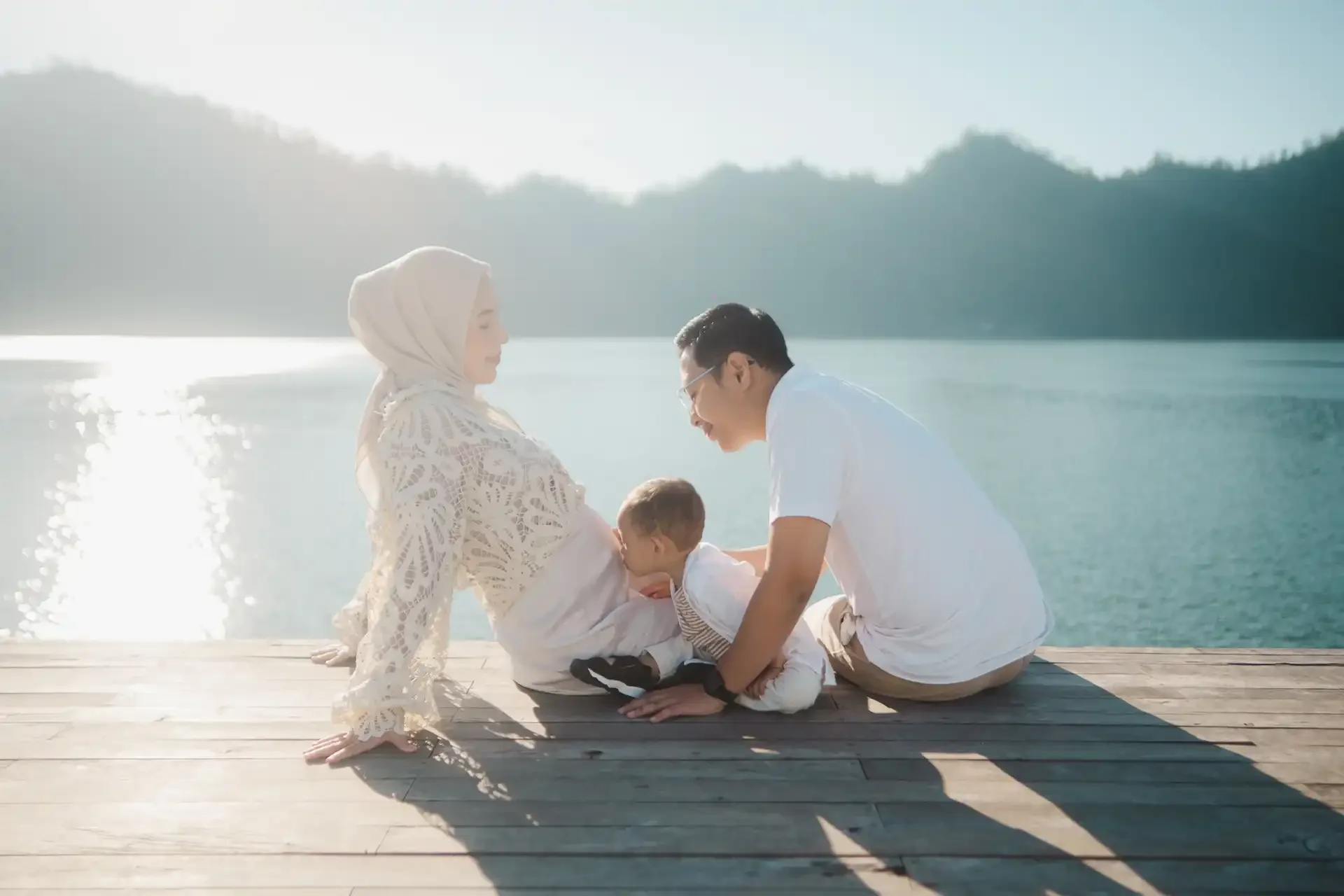 A family sitting on a wooden dock by the water, with a woman in a hijab, a man, and a young child, engaging in a playful and loving moment during sunset or sunrise.