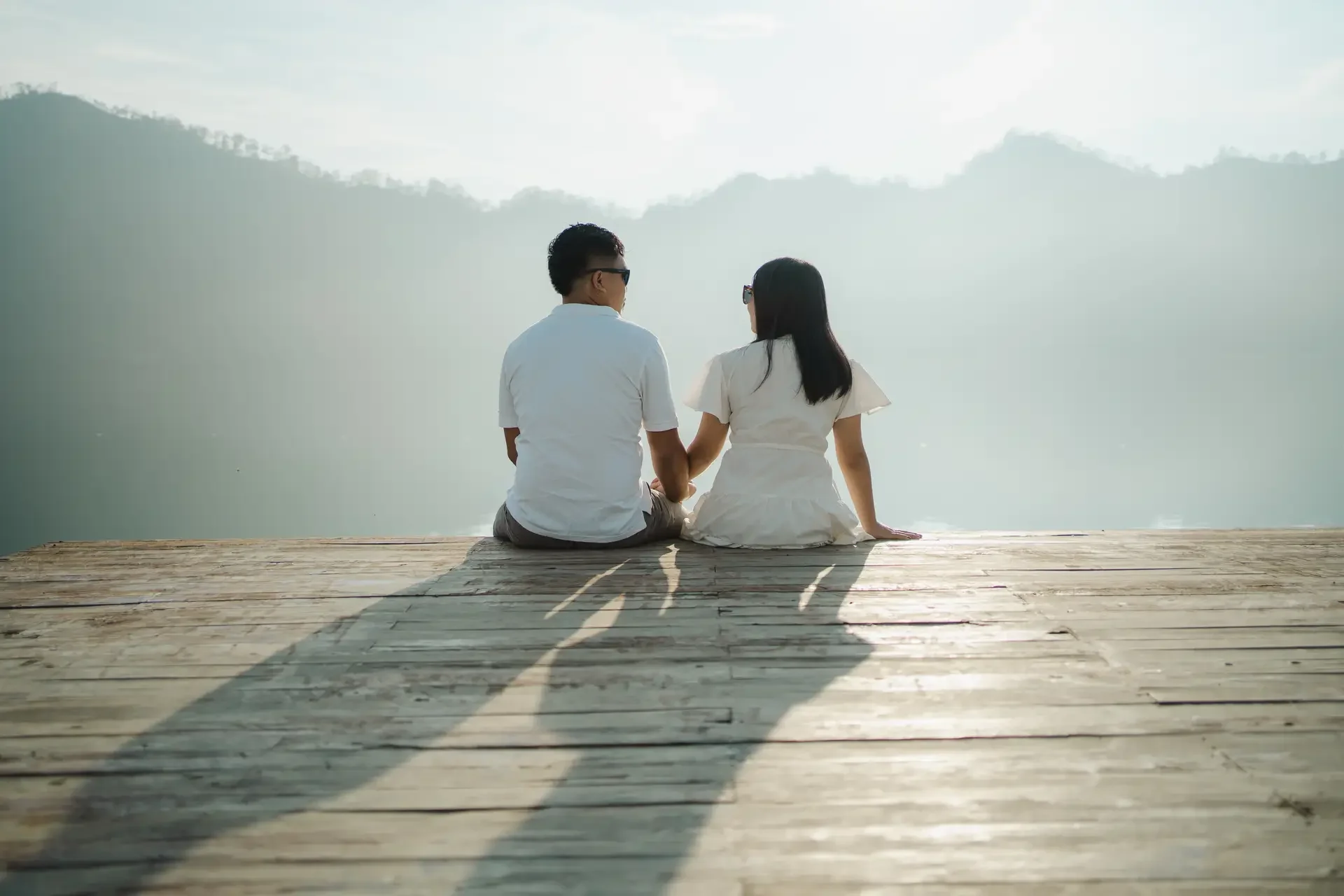 A couple sitting on a wooden dock by a lake, holding hands and facing the water with mountains in the background, during daylight.
