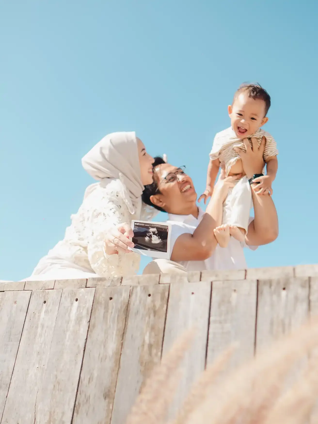 A family enjoying a moment outdoors under a clear blue sky, with a woman in a white hijab and a man holding a young child, who is smiling and wearing a beige striped shirt. The man is holding an ultrasound picture, indicating a joyful family moment.