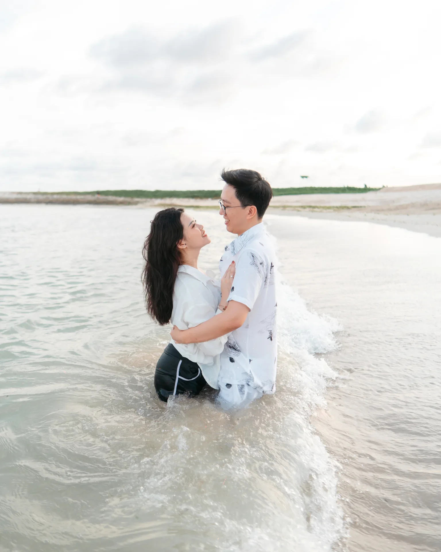 Couple smiling and holding each other in shallow ocean water, embracing on the beach during cloudy day.