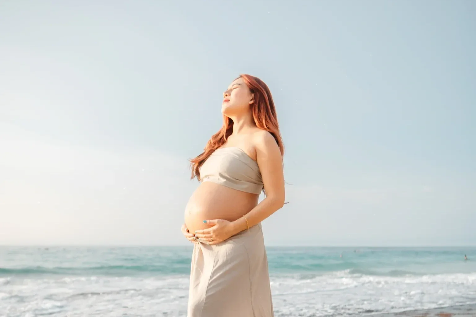 Pregnant woman standing on the beach with eyes closed, facing the sun, wearing a beige strapless top and skirt, with ocean waves in the background.