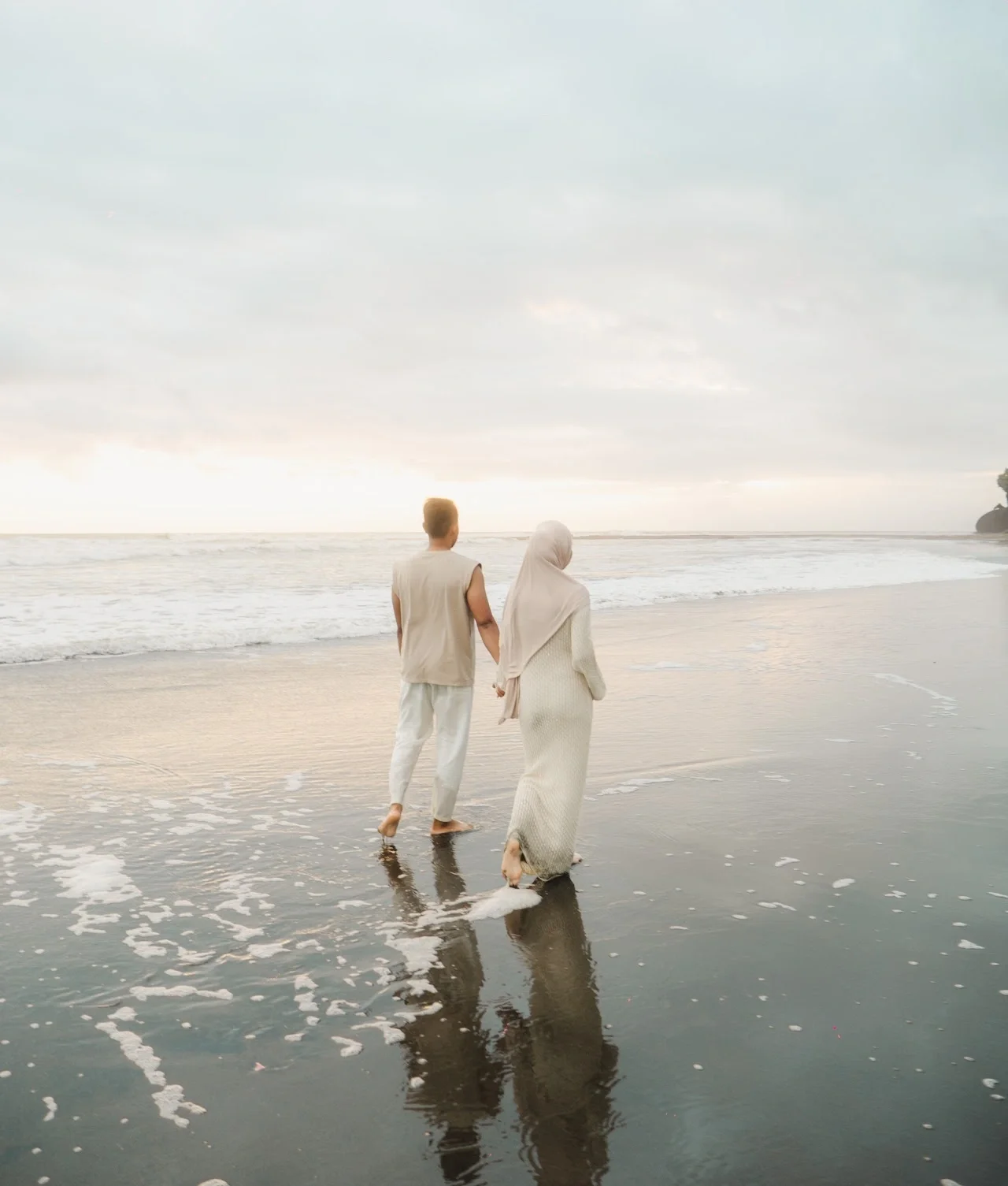 A man and a woman walking hand in hand on the beach near the water's edge, with gentle waves and a cloudy sky overhead.