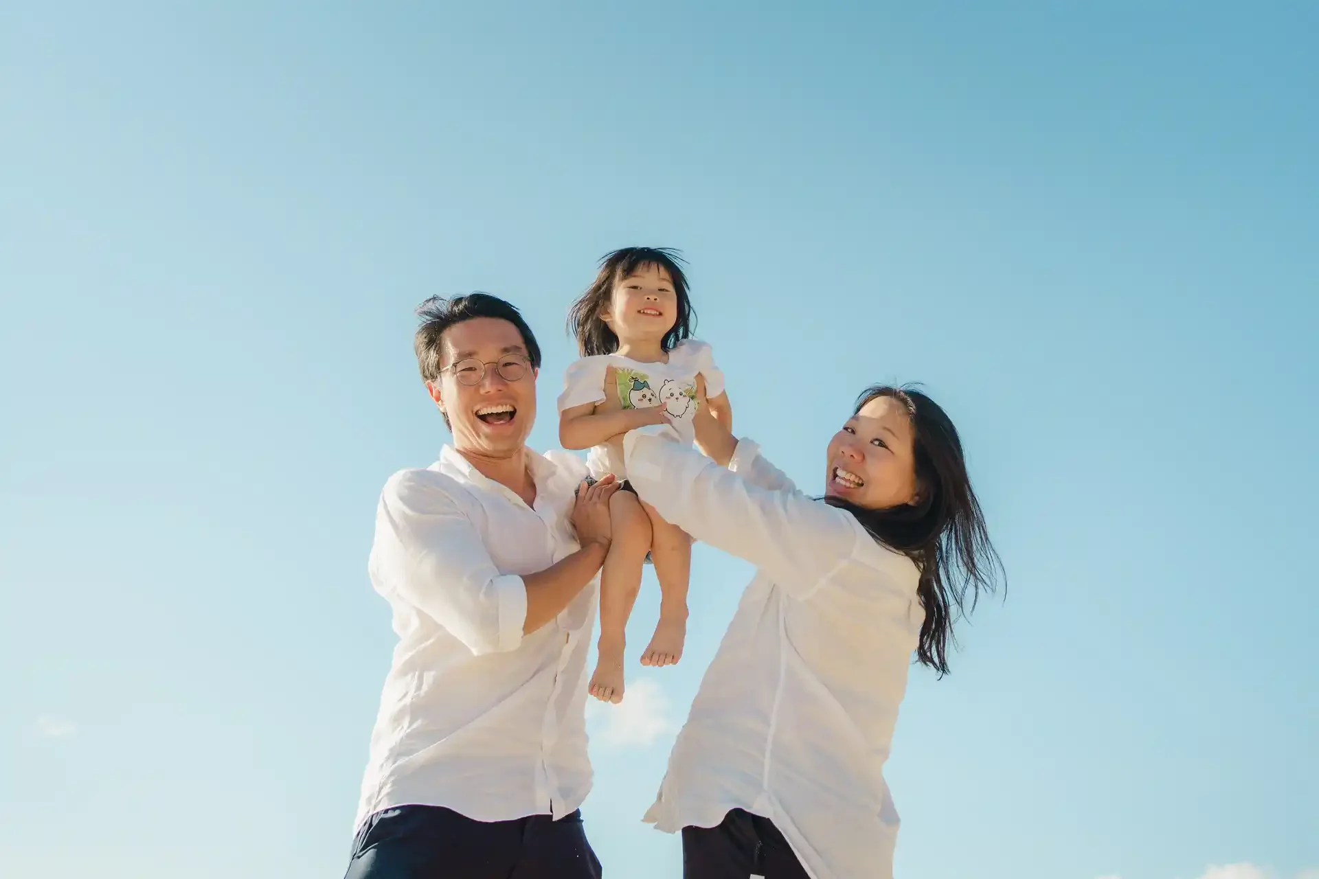 A happy family of three, a man and a woman holding a young girl, enjoying a bright, clear day outdoors with a blue sky in the background.