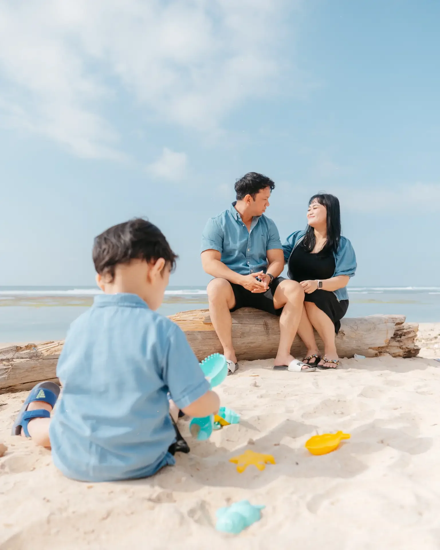 A family at the beach with a young boy playing in the sand and two adults sitting on a log, smiling at each other under a blue sky.