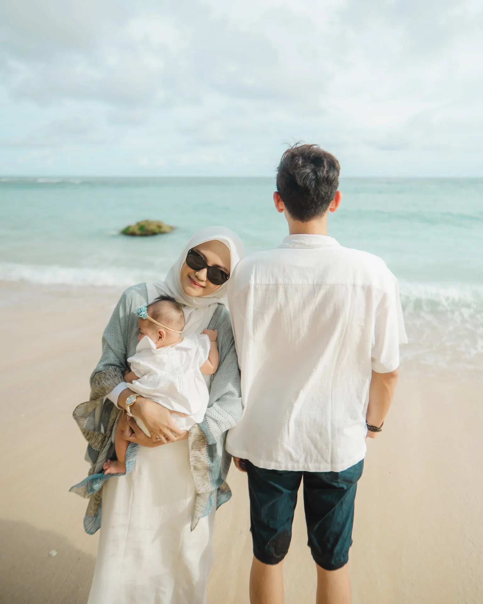 A family at the beach includes a woman wearing sunglasses holding a baby dressed in white, and a man standing with his back towards the camera, facing the ocean.