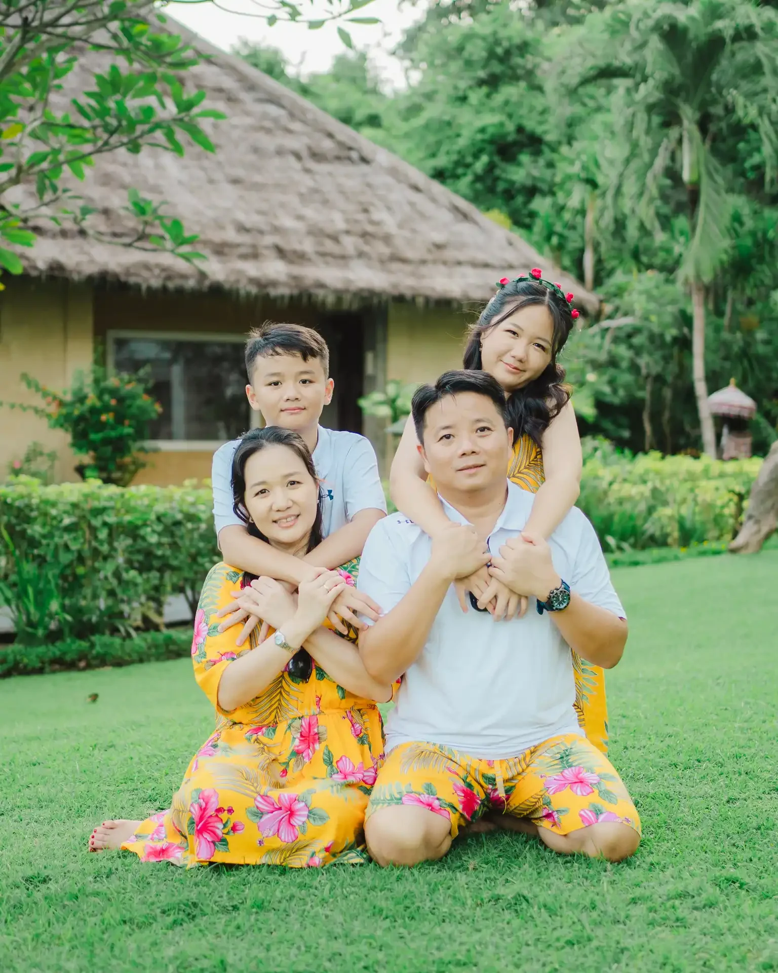 A family of five posing outdoors in a lush garden, dressed in matching yellow floral outfits, with two children seated on their parents' shoulders and the parents kneeling on the grass.