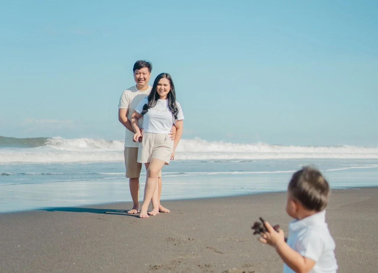 Foto keluarga romantis di pantai Bali dengan ombak dan langit mendung dramatis.