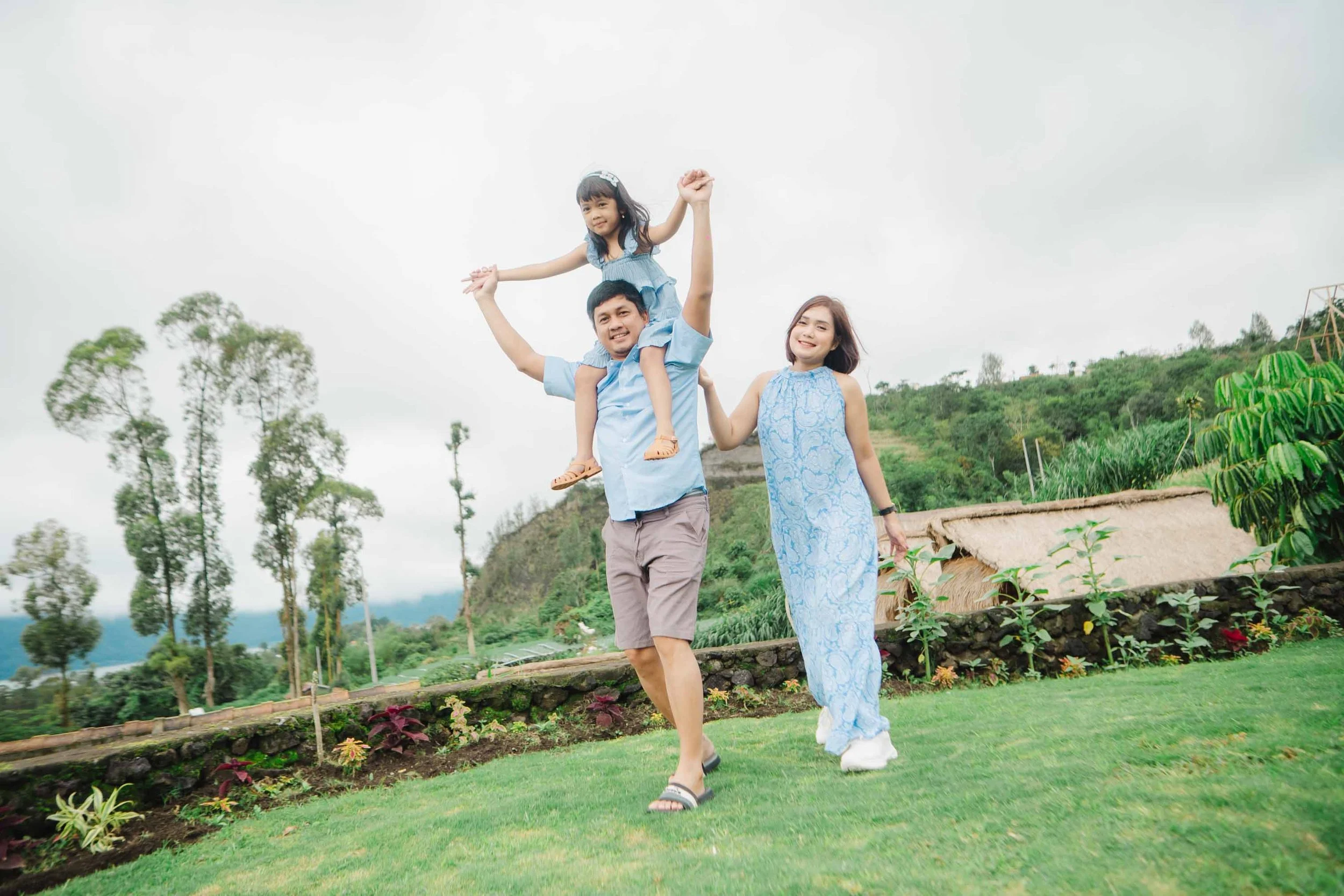 A family of three, a man, a woman, and a young girl, is enjoying time outdoors on a grassy area with trees and a hill in the background. The man is carrying the girl on his shoulders, while the woman is holding his hand and smiling.