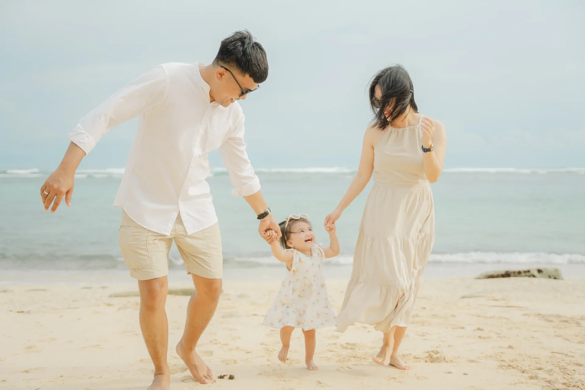 A family of three, a man, a woman, and a young girl, enjoying a day at the beach. The man and woman are holding the girl’s hands as they walk on the sand, smiling and playing. The ocean is in the background with waves and an overcast sky.