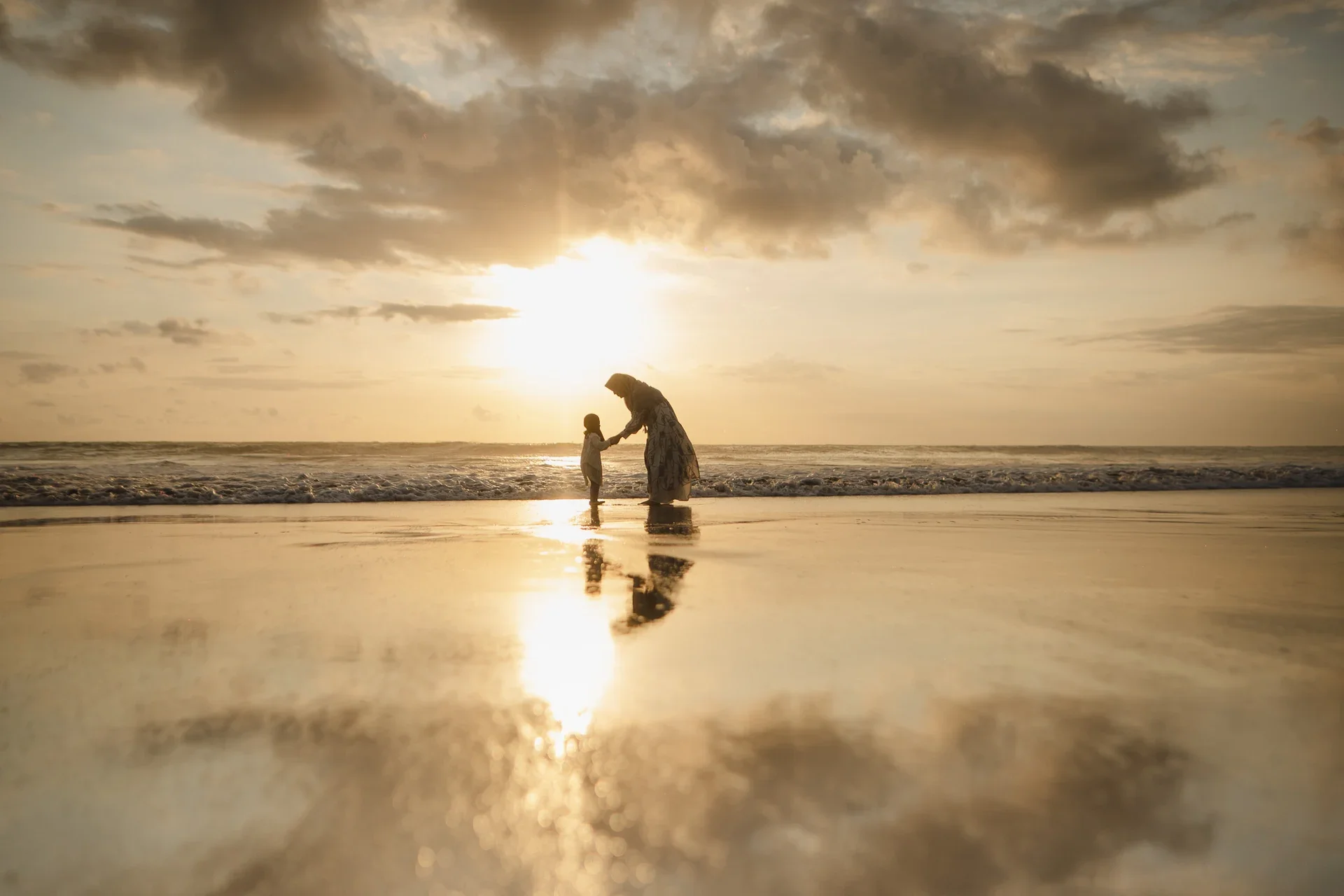 A woman and child standing on the beach at sunset, holding hands, with their reflections visible on the wet sand.