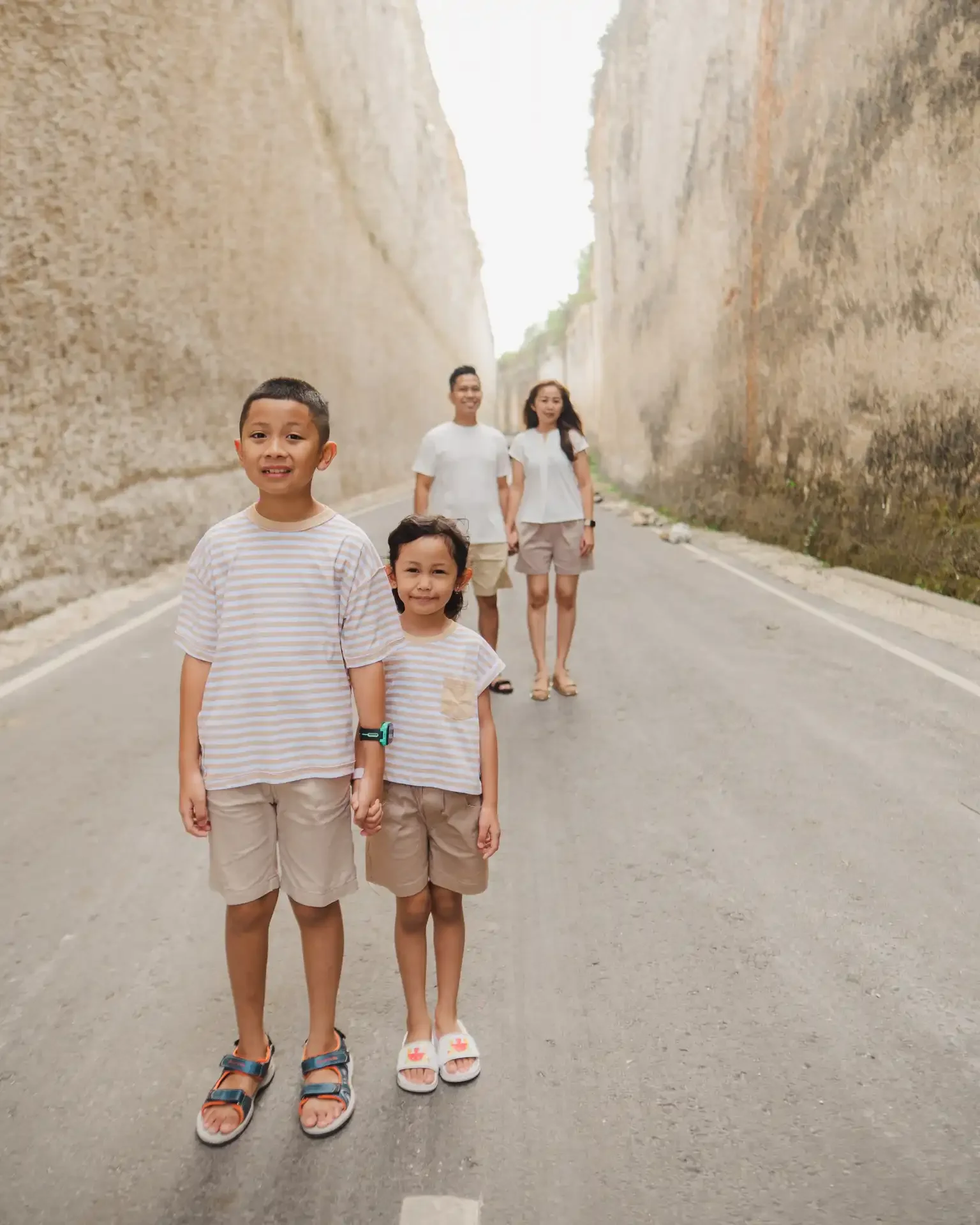 A family of five standing on a road between tall, rocky cliffs. Two children are in the foreground, holding hands, with three adults in the background.