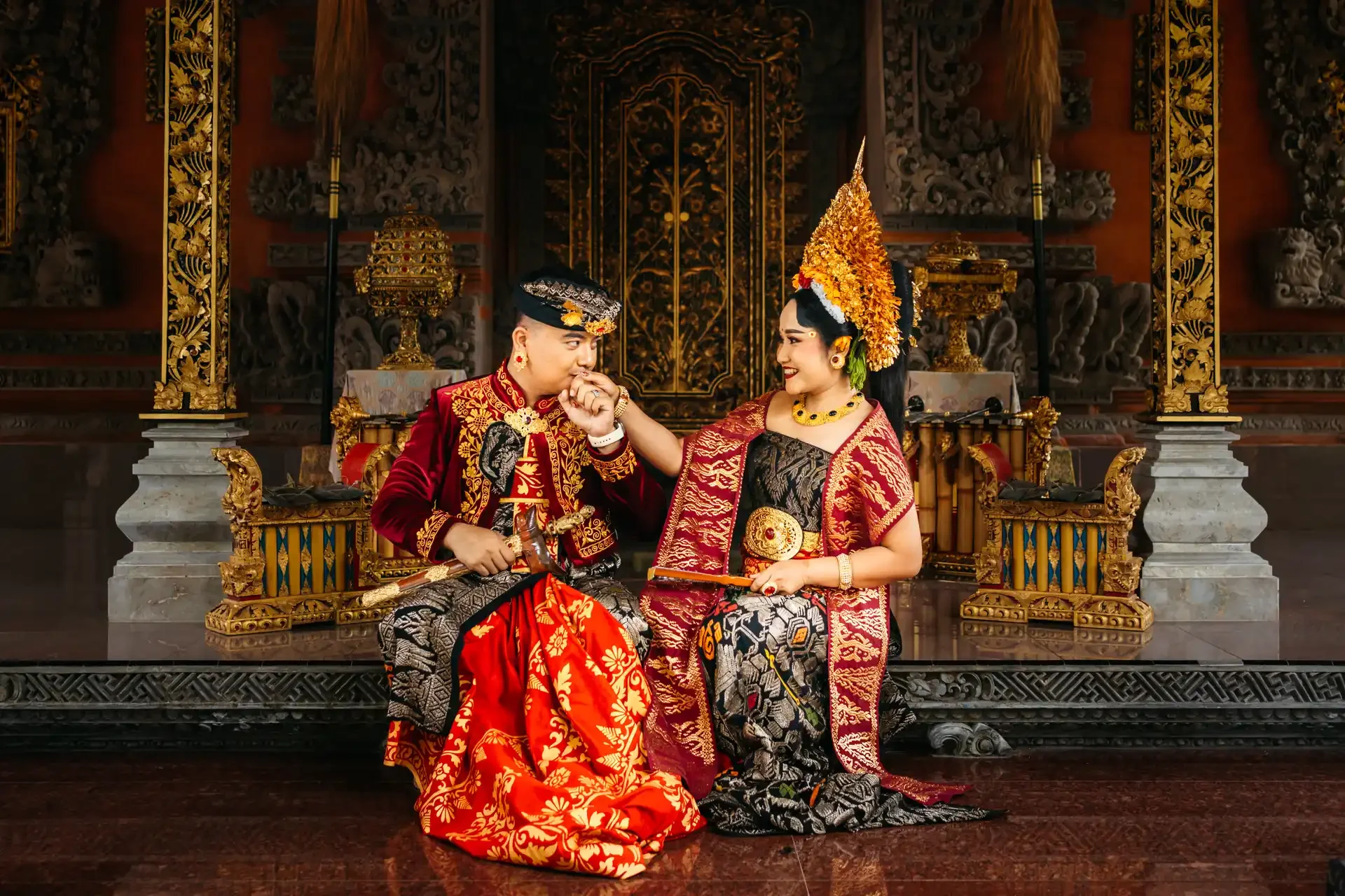 A couple wearing traditional Indonesian wedding attire sitting on a regal throne in a decorated hall.