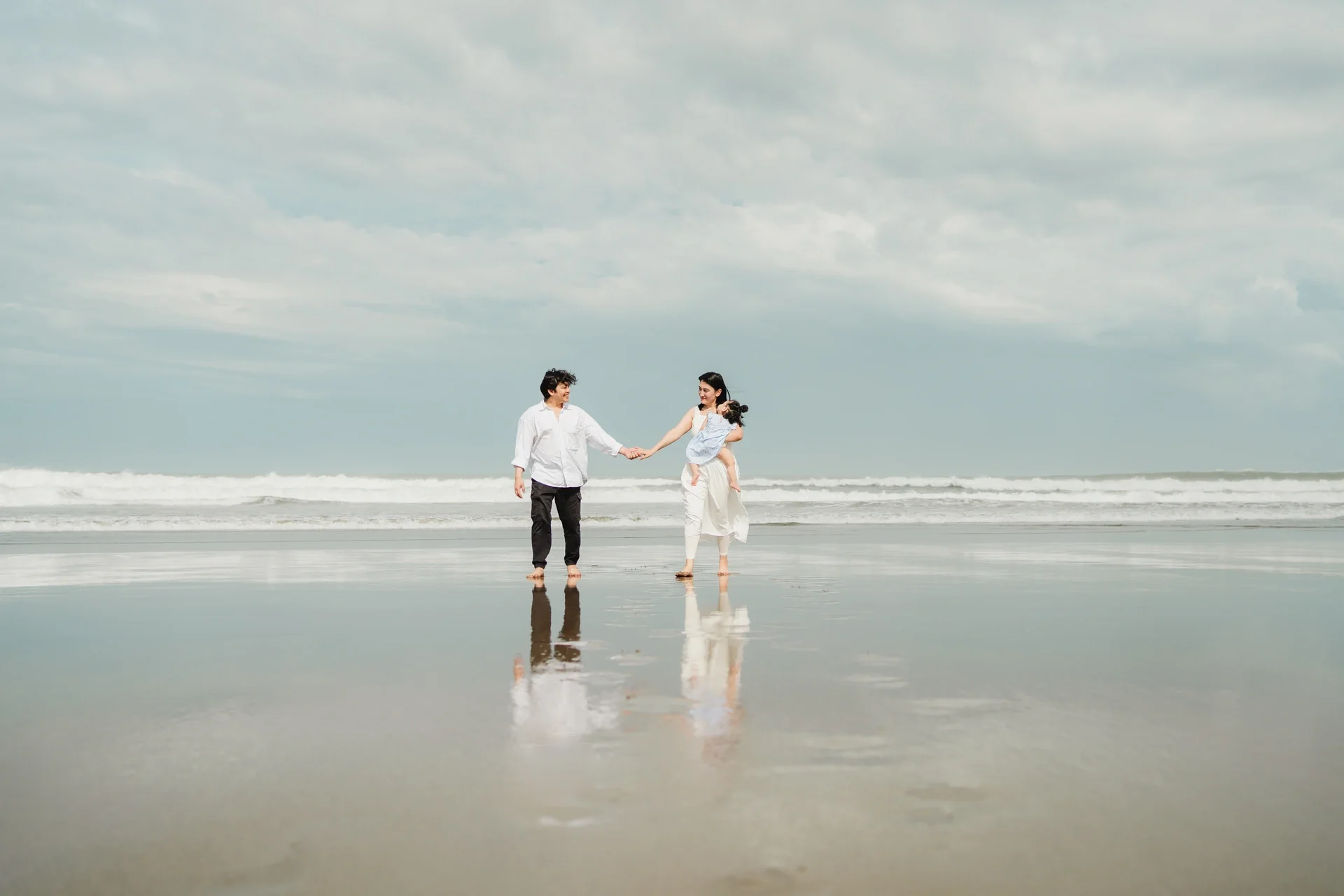 A family of three walking on the beach, holding hands, with the ocean and cloudy sky in the background.