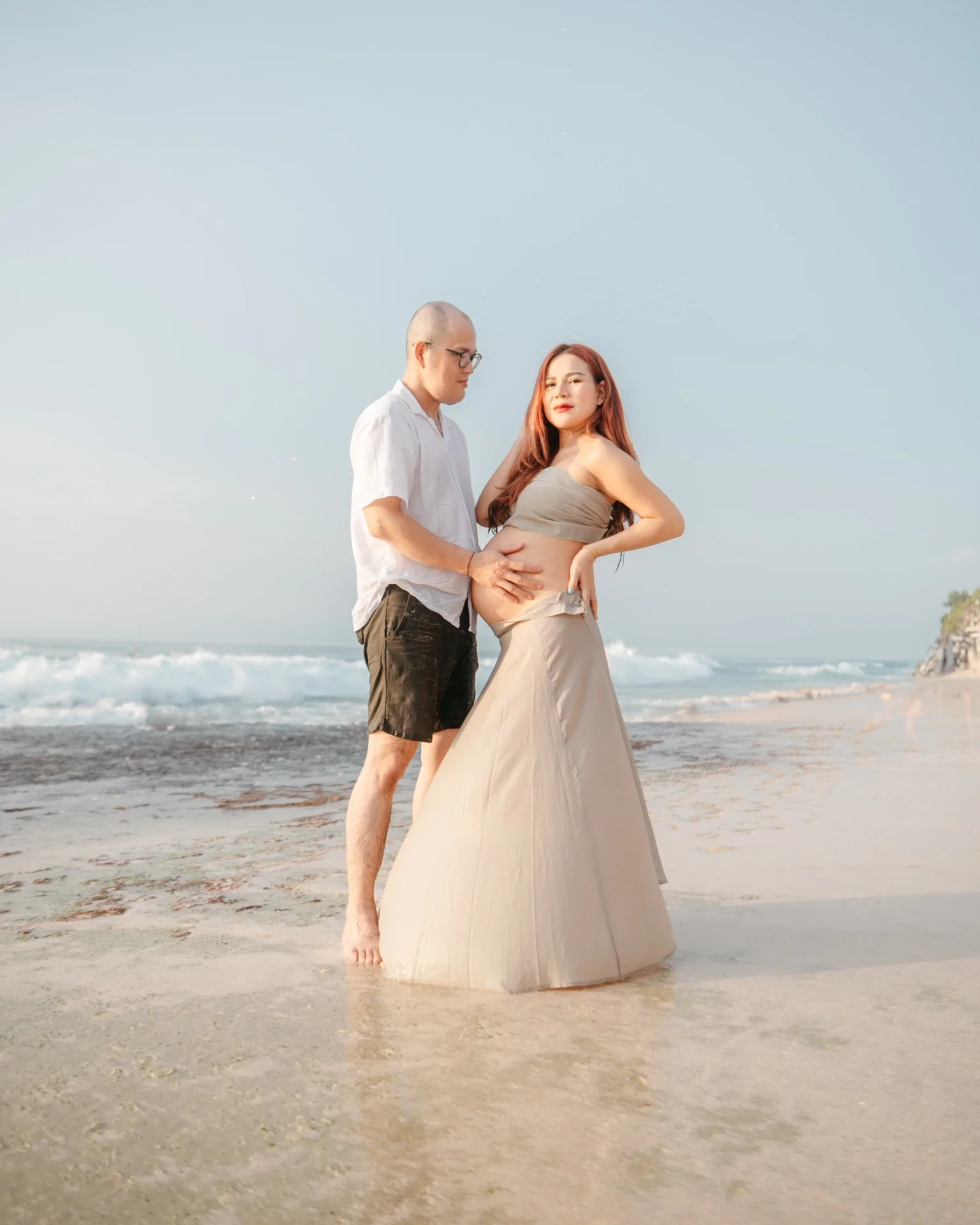 A man and a pregnant woman standing on a beach with ocean waves in the background. The man is holding the woman's belly while she looks at the camera. The woman is wearing a beige dress, and the man is dressed in a white shirt and dark shorts.