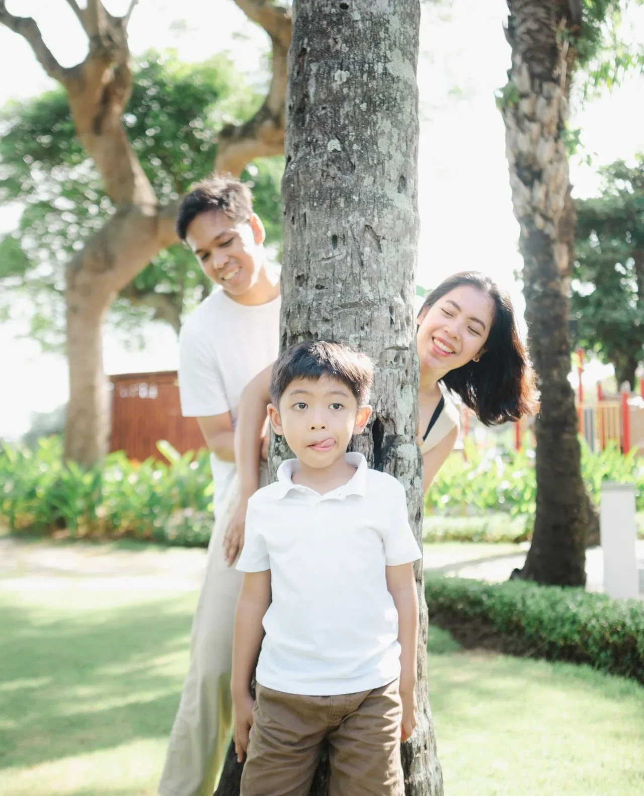 A family of three playing hide and seek outdoors behind a tree in a park with green grass and trees.