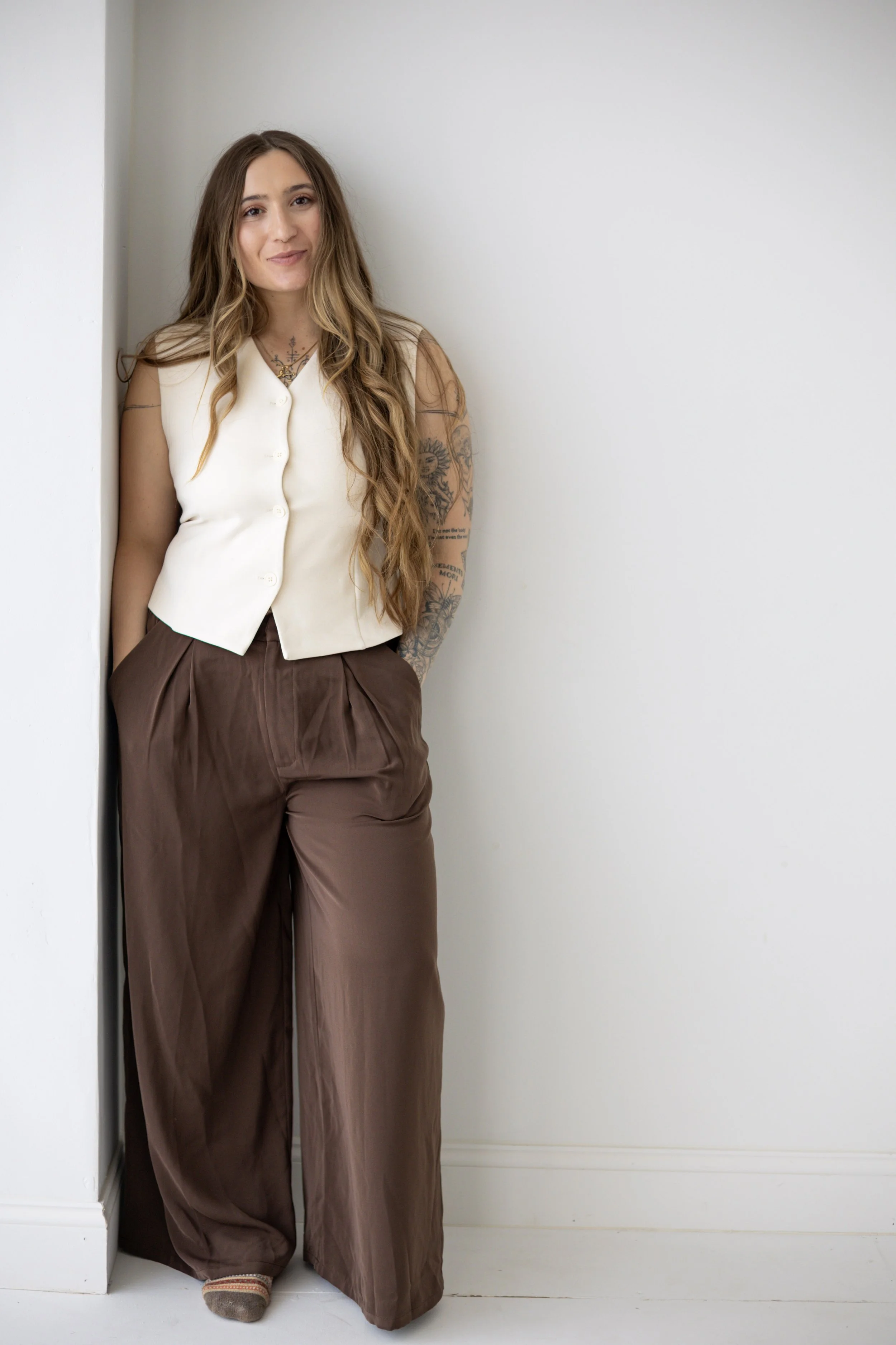A young woman with long wavy hair, wearing a sleeveless cream blouse and brown wide-leg pants, standing in a corner against a plain white wall. She has tattoos on her left arm and neck, and is smiling softly.