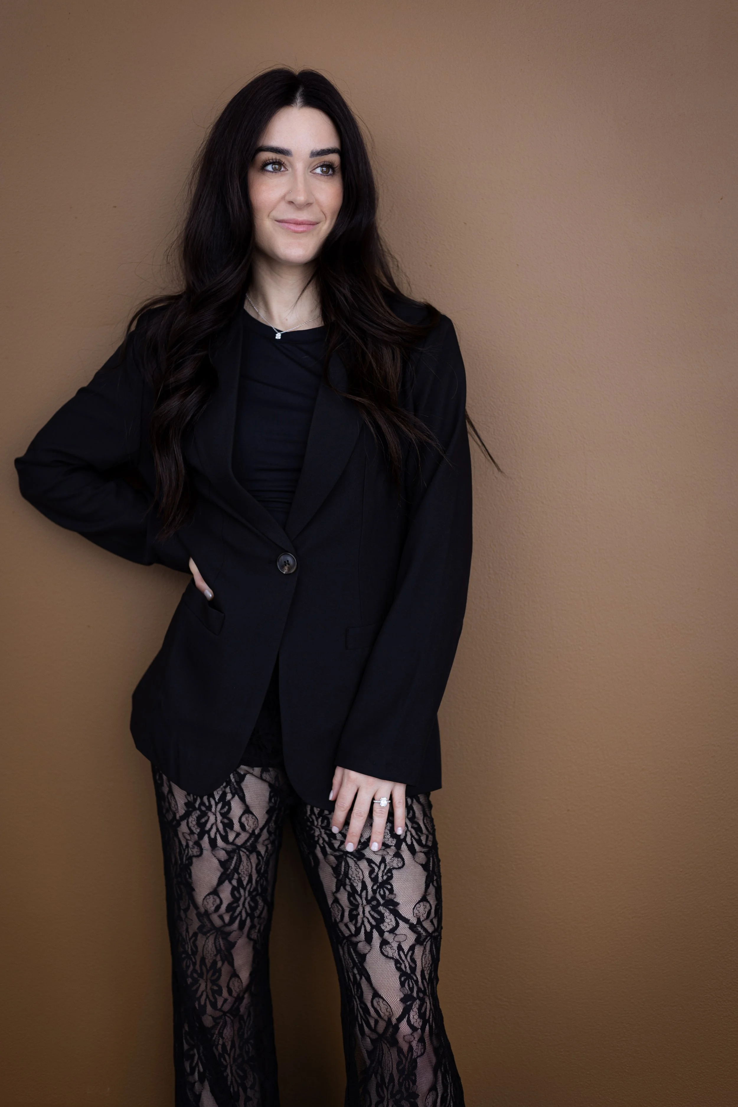 A woman with long dark hair wearing a black blazer and matching lace pants standing against a plain brown wall.