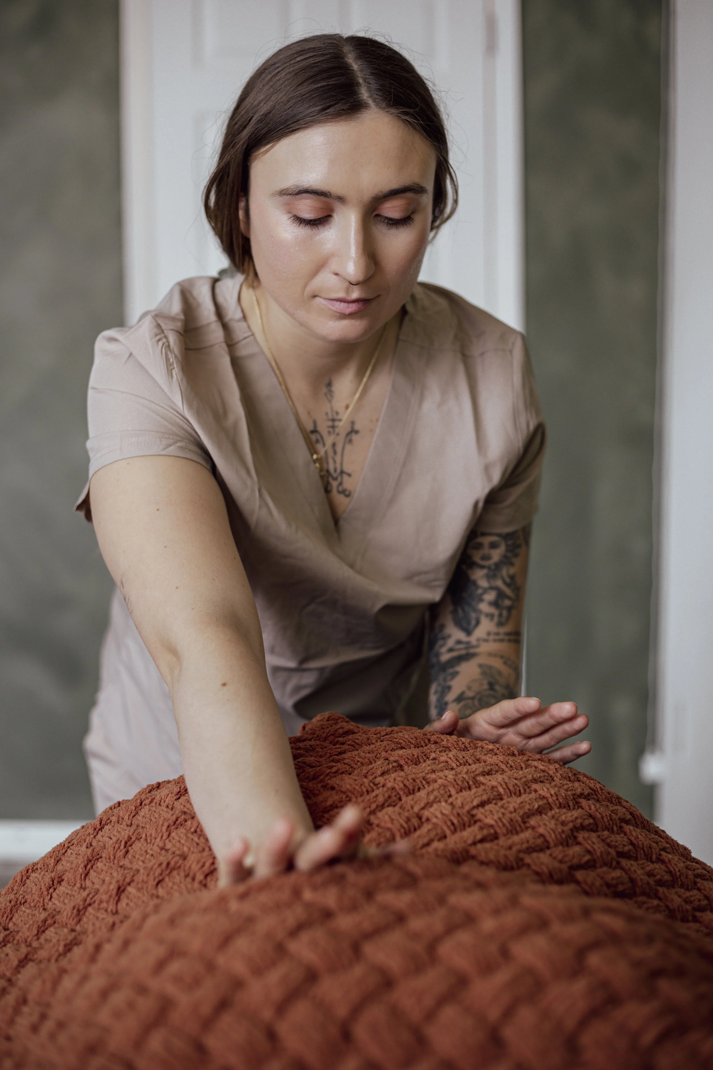 A woman with short brown hair and tattoos on her arms is leaning over and pressing her hands on a textured, reddish-brown blanket or fabric.
