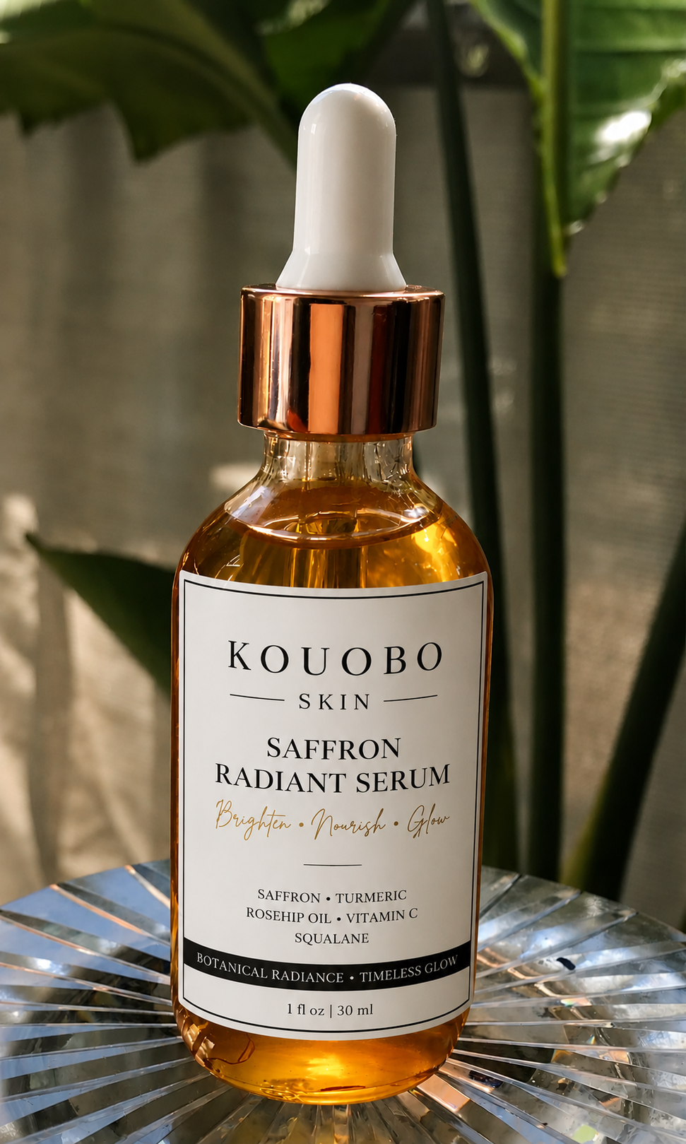 A bottle of Koubo Skin Saffron Radiant Serum with a white dropper cap, placed on a reflective metallic surface with a leafy green plant in the background.