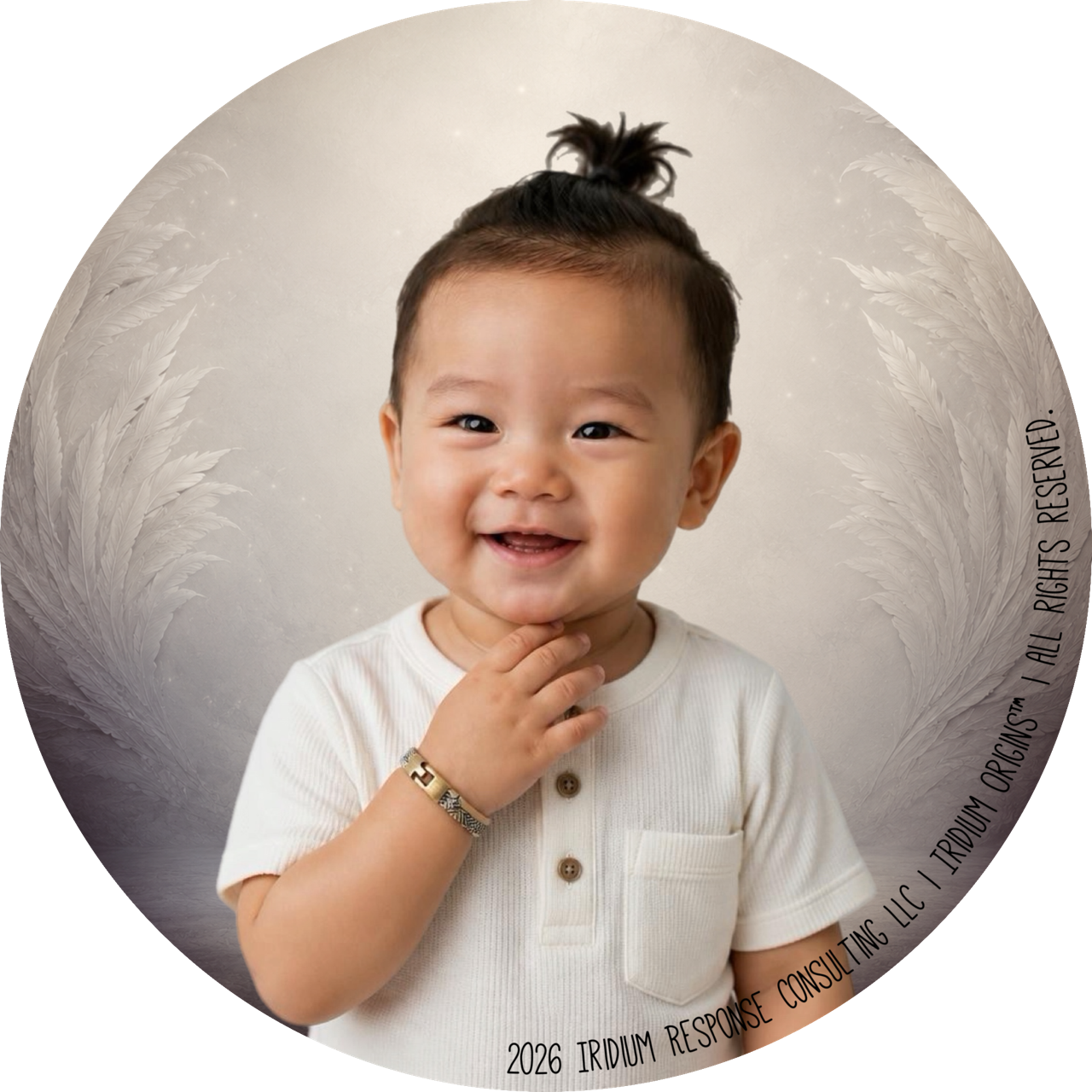 A smiling young boy with short black hair styled in a small top knot, wearing a white shirt with a front pocket and brown buttons, holding his chin with one hand and wearing a gold bracelet, standing in front of a light-colored background with feathers and sparkles.