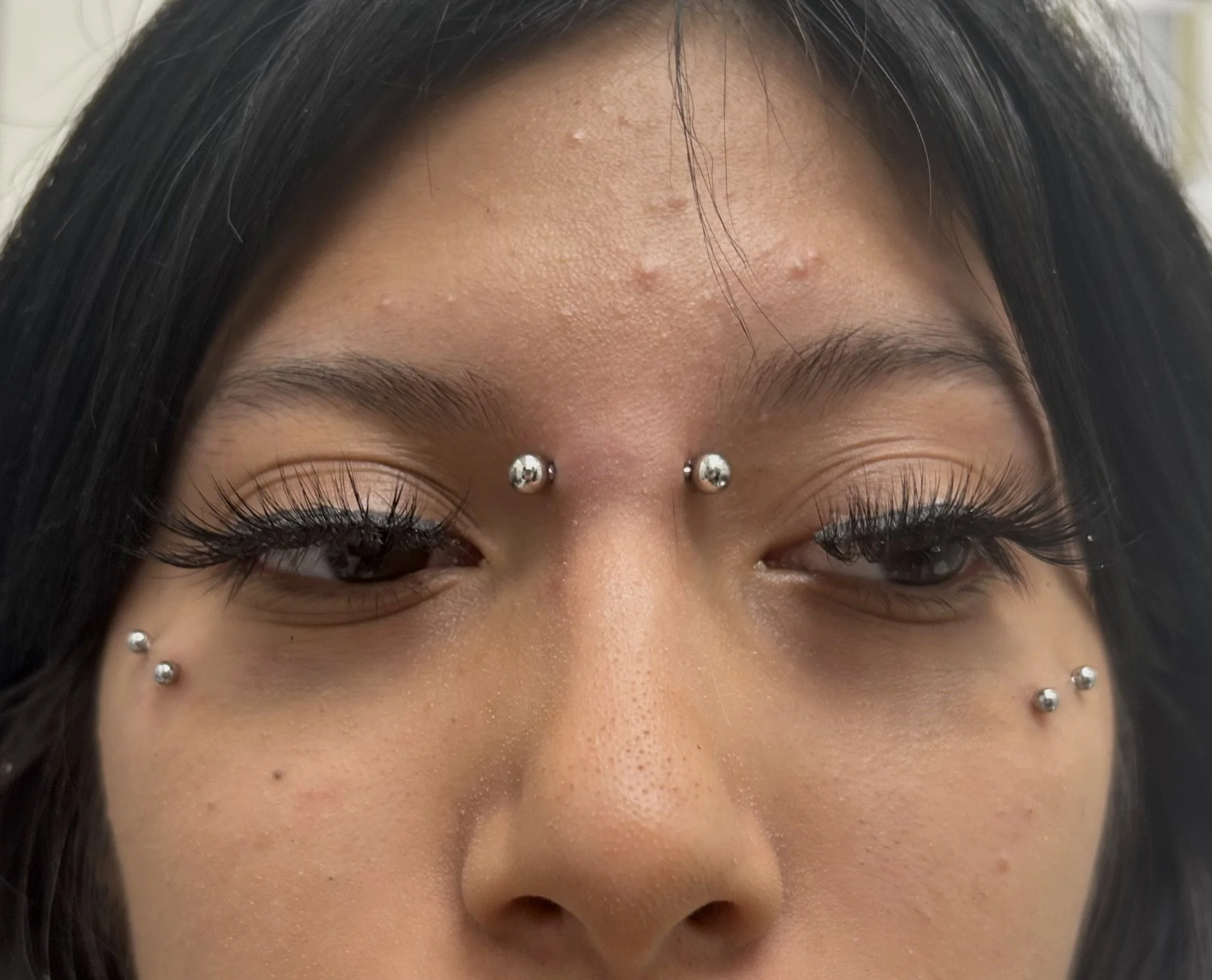 Close-up of a woman's forehead with pimple scars and jewelry piercings around the eyebrows. She has dark hair and long eyelashes.