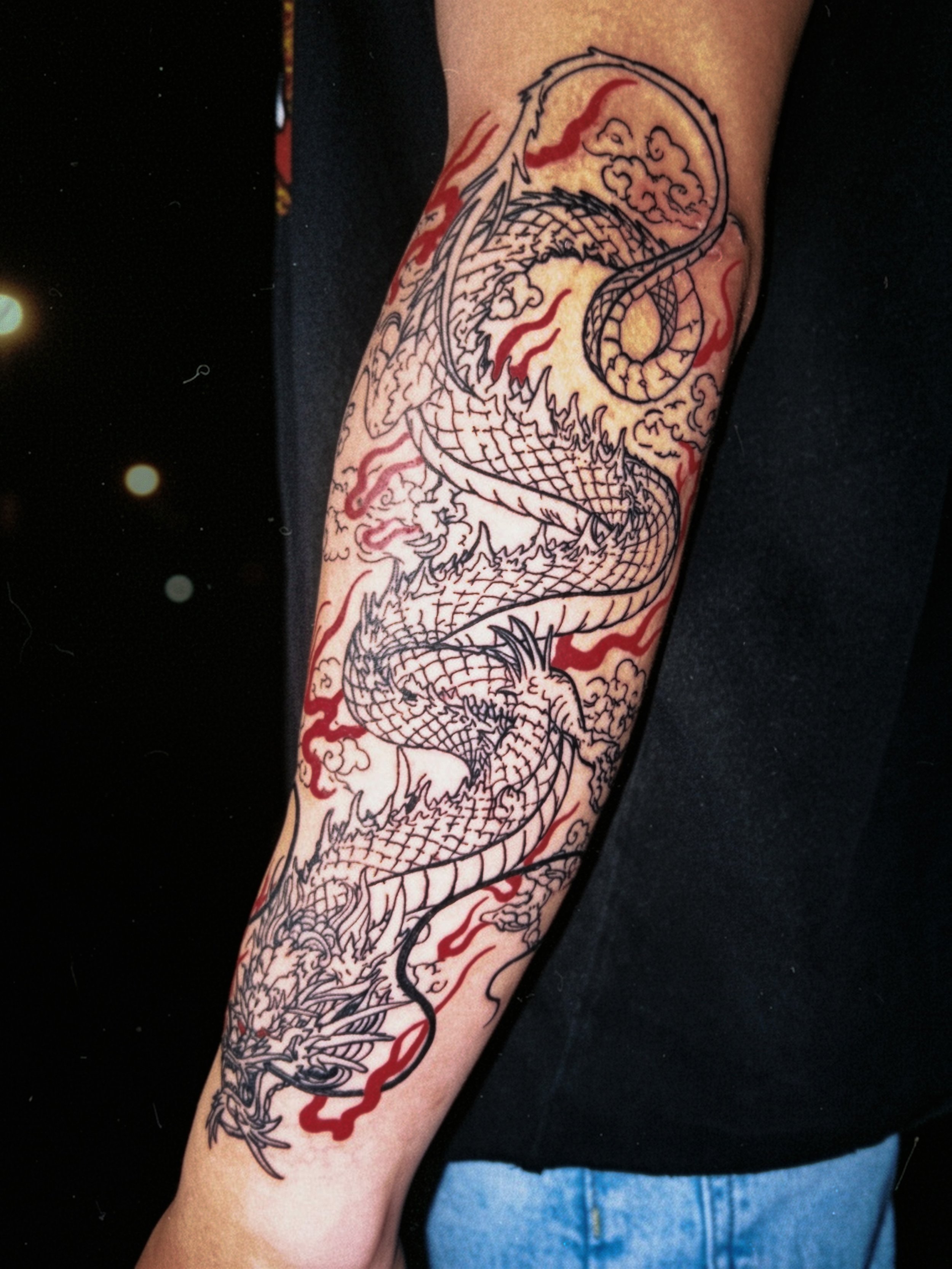 Tattoo of a dragon on a person's forearm, with detailed scales and flames, in black and red ink.