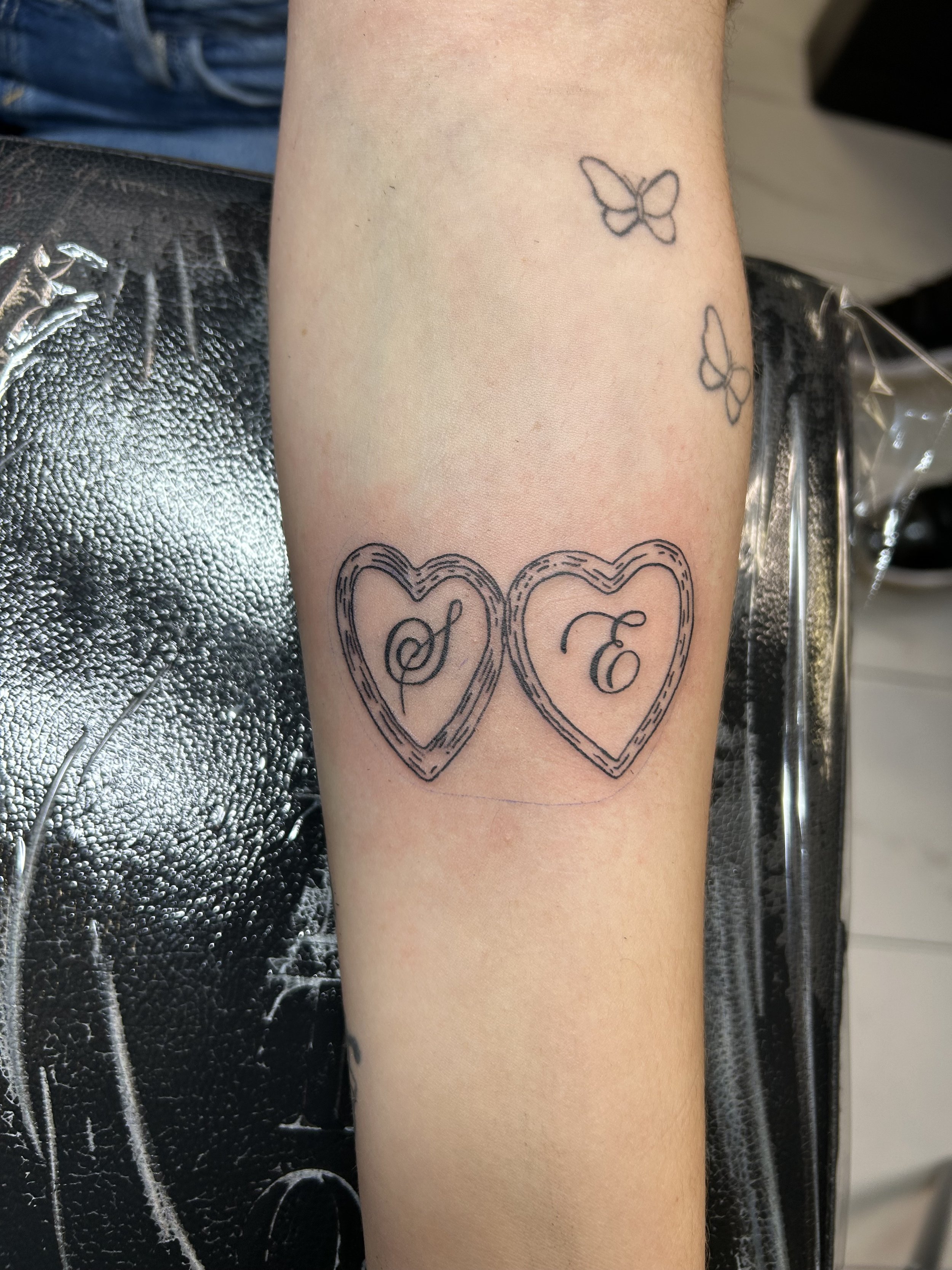 Tattoo on forearm of two overlapping hearts with initials inside, and three butterfly outlines above it.