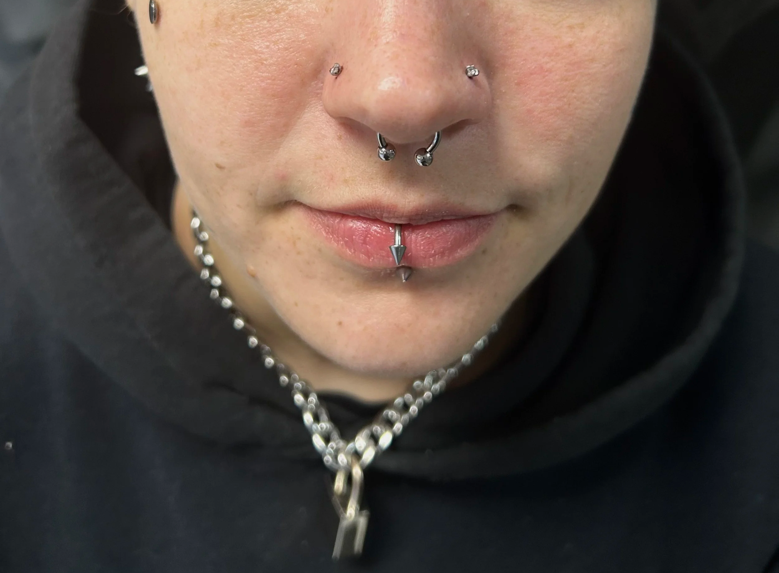 Close-up of a person's face showing multiple facial piercings, including a septum piercing, two nostril piercings, a labret piercing, and a piercing on the lower lip, with a chain necklace visible.