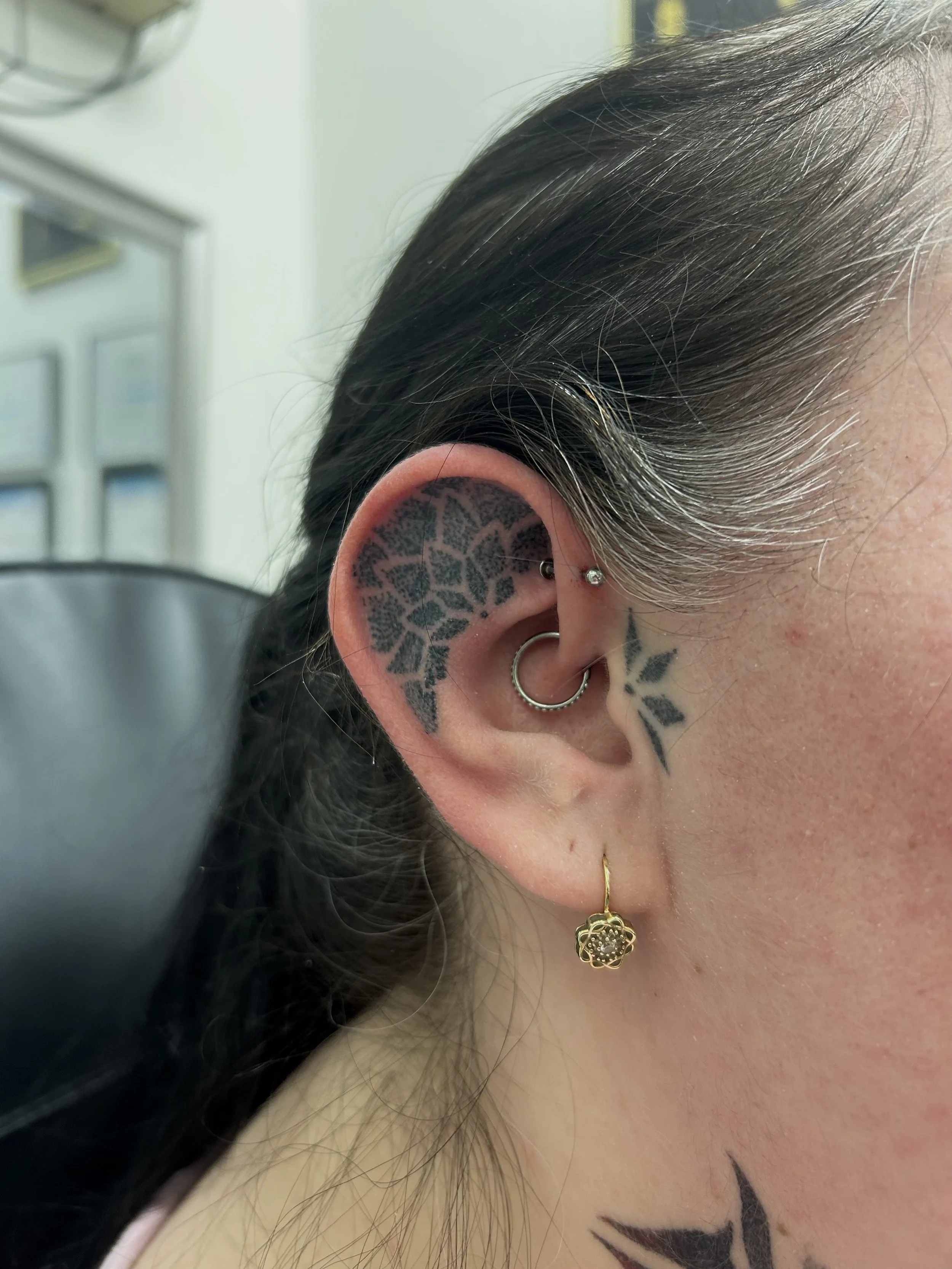 Close-up of a person's left ear with multiple earrings, includes black and gray tattoos behind the ear, dark brown hair, and a gold earring hanging from the earlobe.