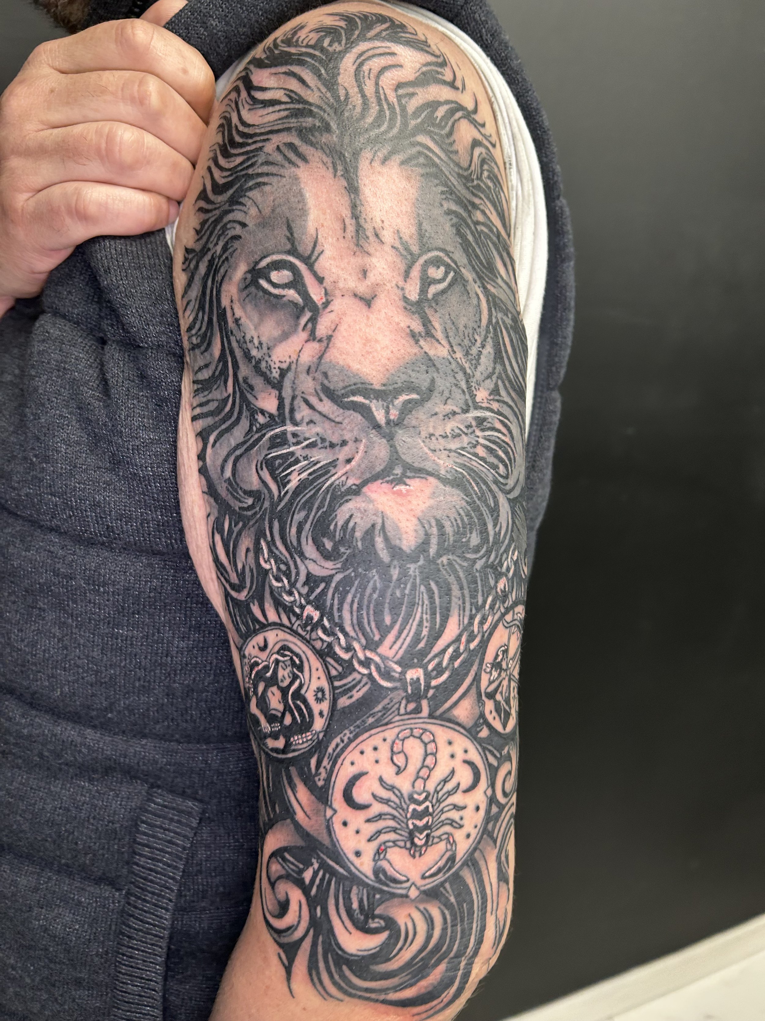 Tattoo of a lion's face on person's upper arm with detailed mane, surrounded by scorpion and fortune-telling symbols.