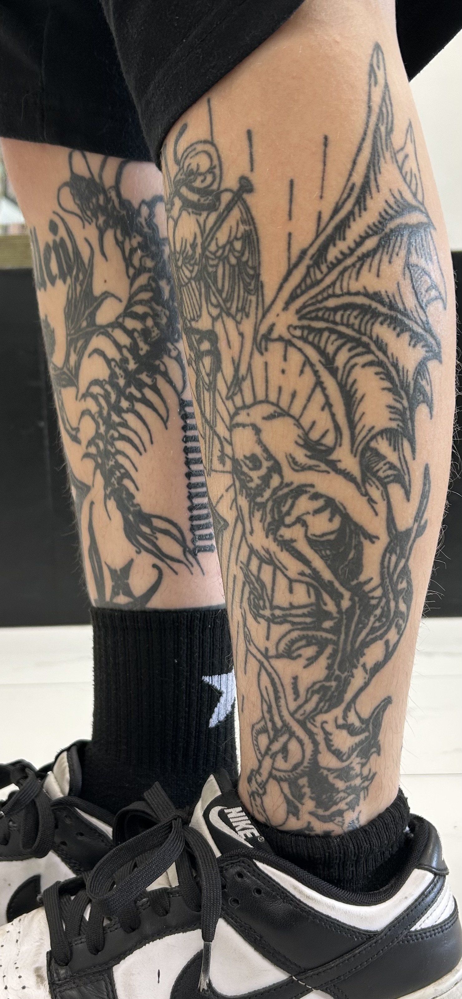 Tattoo of a winged demon or lion with sharp claws, a humanoid figure with wings and a spear, and abstract designs on a person's leg, wearing black and white Nike sneakers and black socks.