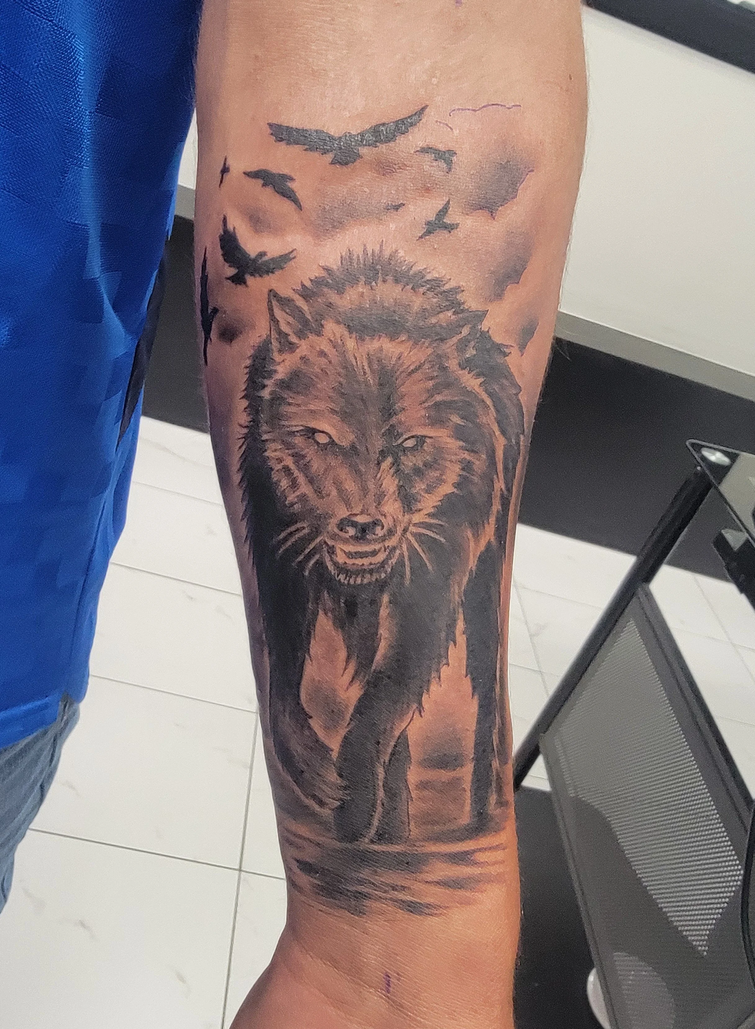 Tattoo of a wolf on a person's arm, with a background of flying birds and clouds.