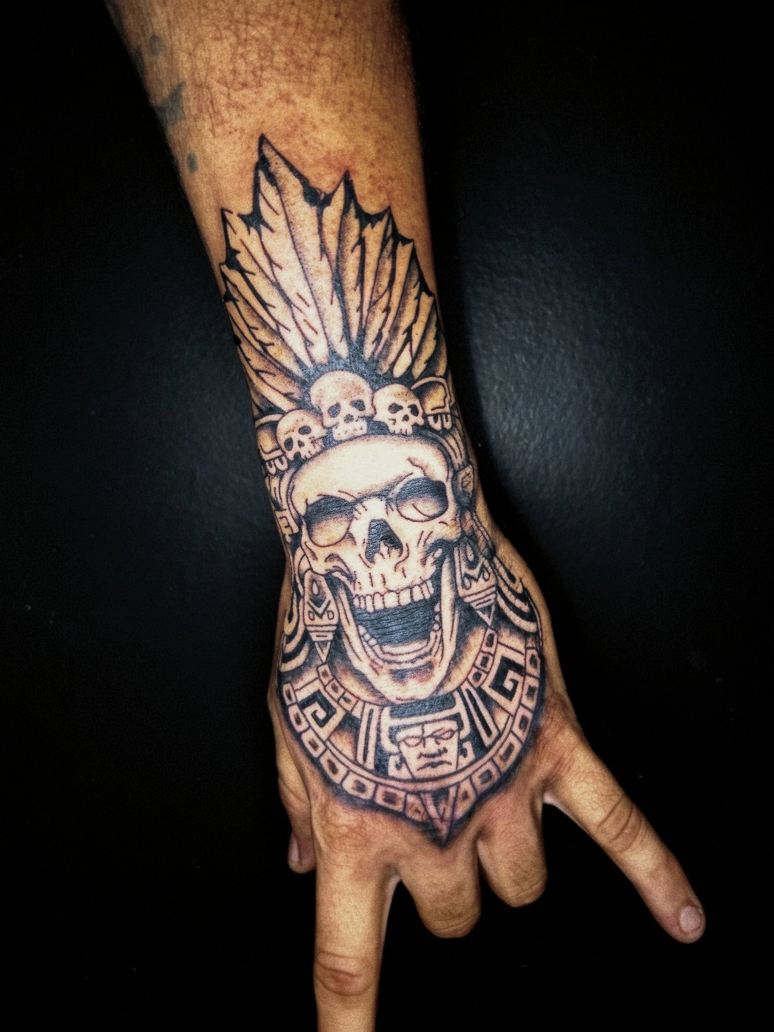 Tattoo of a skull wearing a headdress with feathers and three smaller skulls above it on a person's forearm.