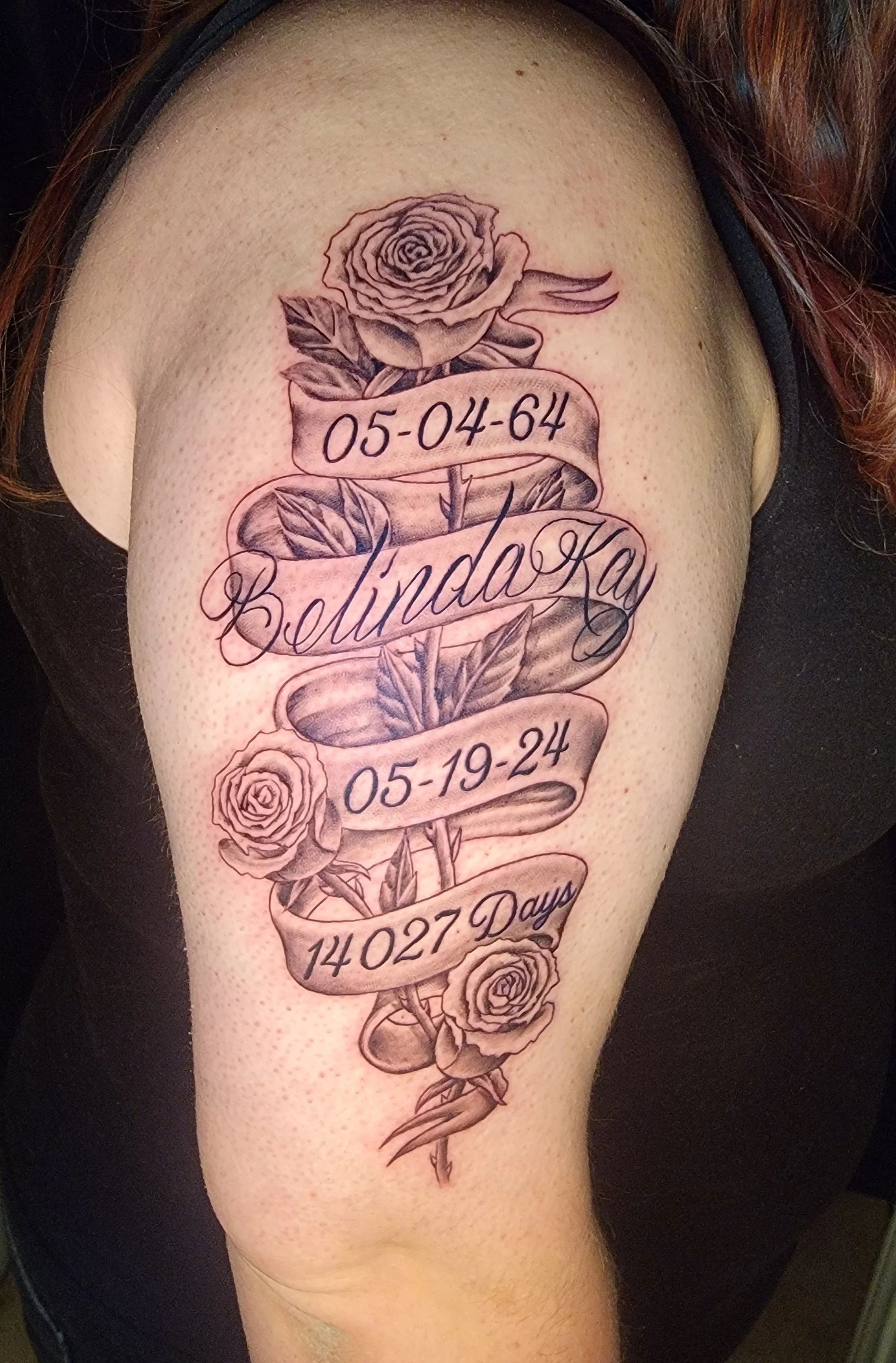 A tattoo on a person's upper arm featuring roses, leaves, and a ribbon with dates, a name, and the phrase '14027 Days' written on it.