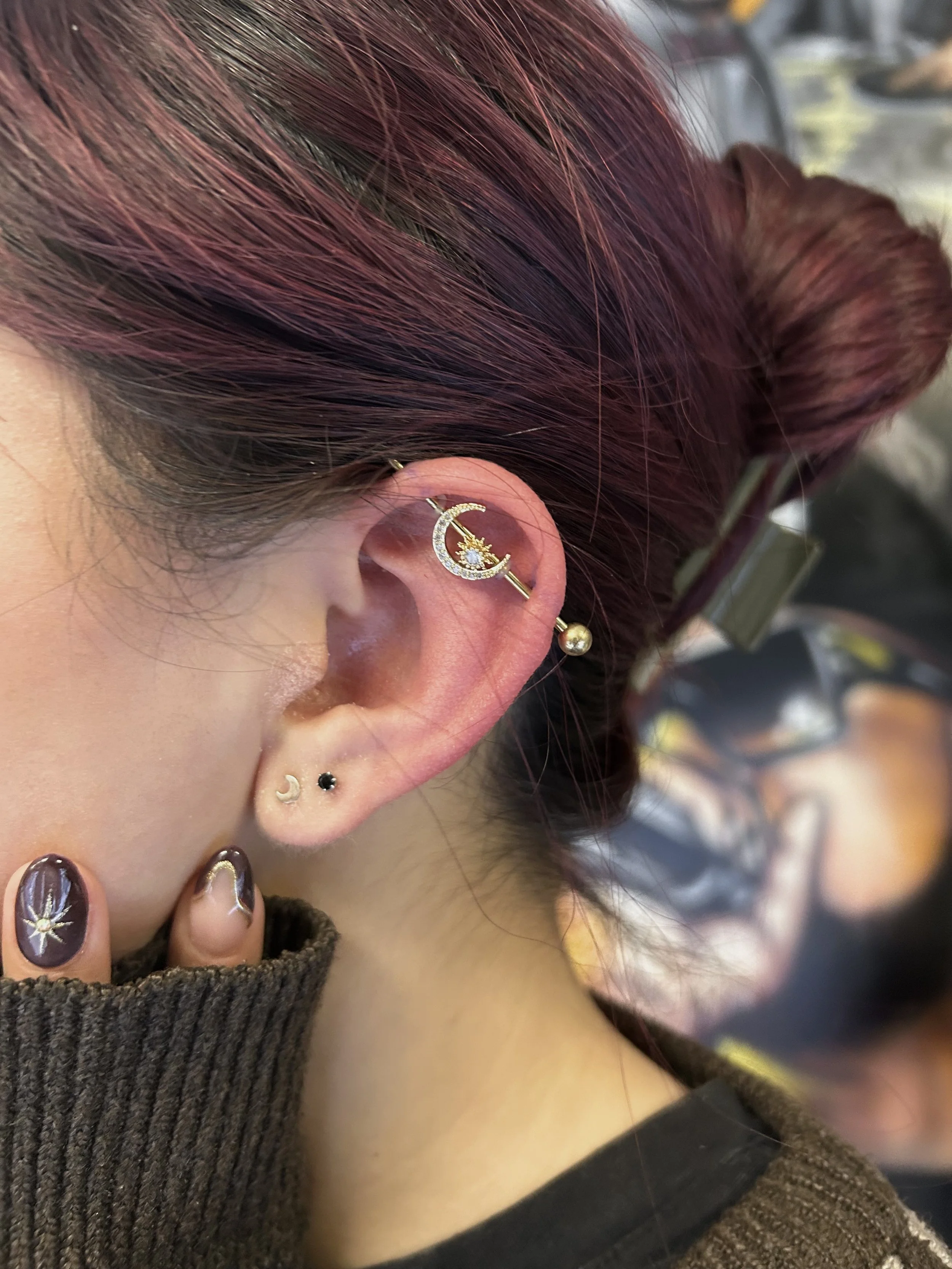 Close-up of a person's ear with multiple earrings, including a moon and star design with the moon outlined in rhinestones, a small black stud, a gold ball, and another small moon. The person has reddish hair styled in a bun and is wearing a brown sweater with a black shirt underneath.