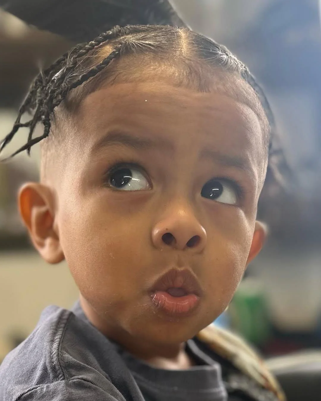 A young child with braided hair and a curious expression, looking to the side indoors.
