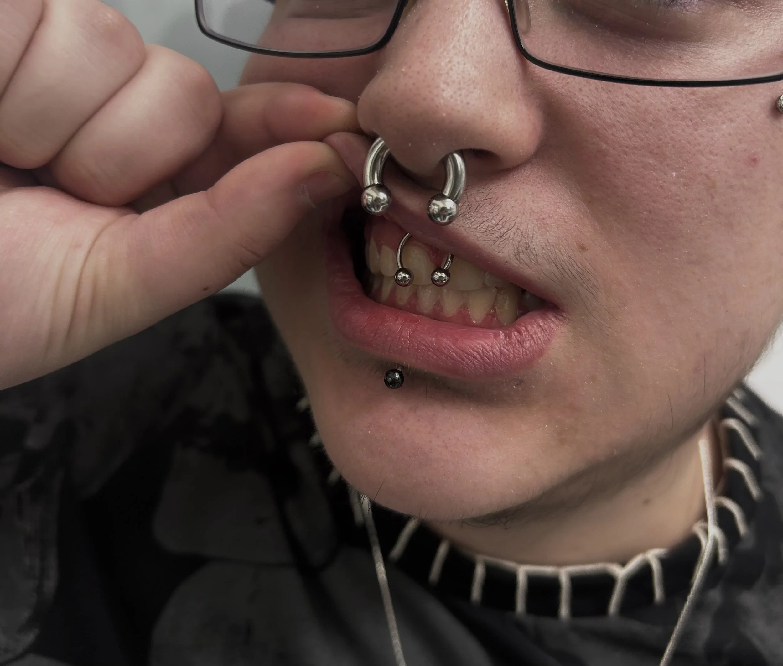 A close-up of a person’s face with light skin and freckles, adjusting their black-rimmed glasses with their hand. The person has multiple facial piercings including a septum ring, a lower lip piercing, and a piercing on the eyebrow, with short facial hair visible on the chin and upper lip.