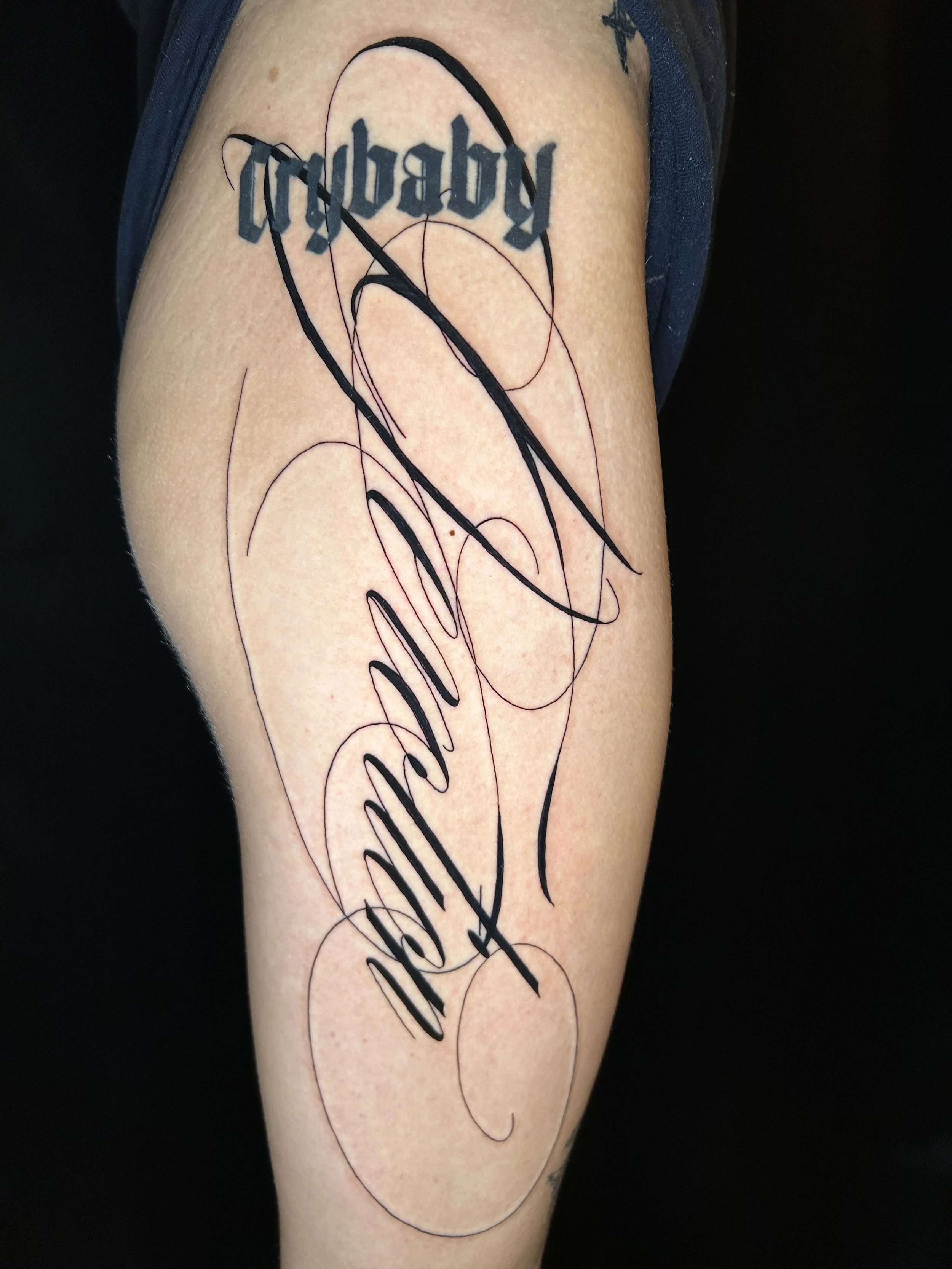 Tattoo on person's upper arm with the words 'Baby Mama' in black cursive, overlapping with abstract black ink lines.