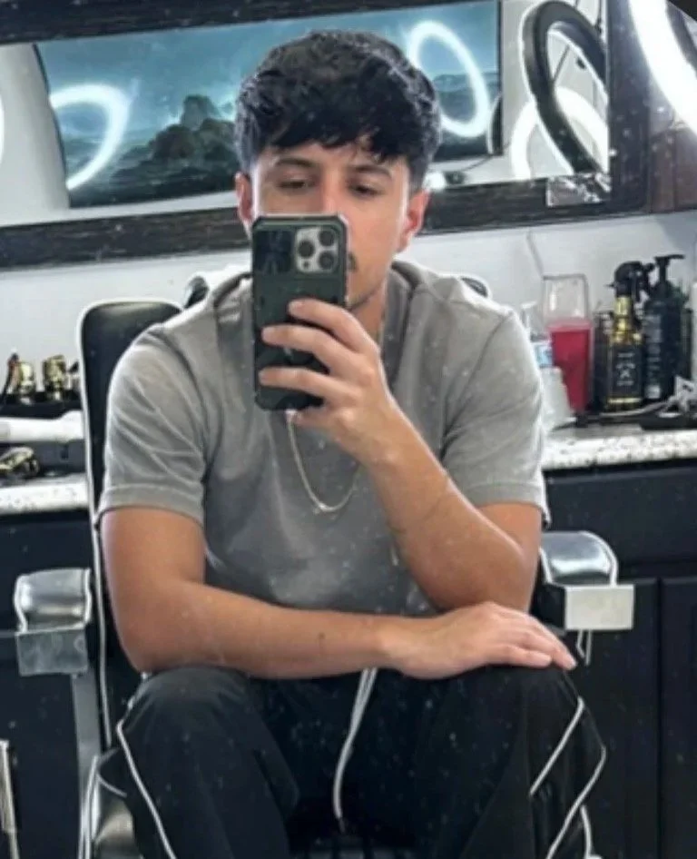 Young man taking a mirror selfie in a barbershop or salon, with various hair products and tools on the counter behind him.