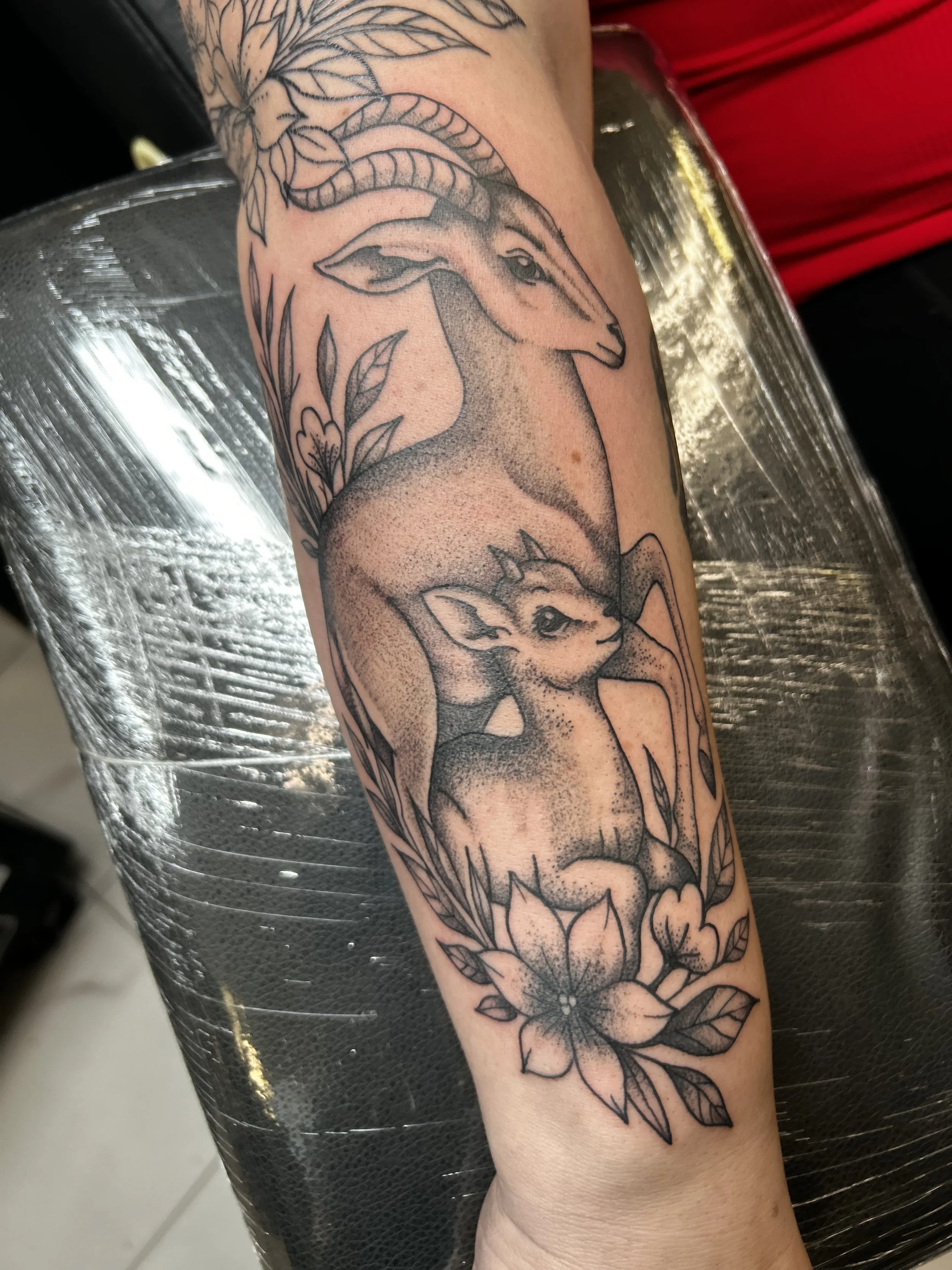 Black outline tattoo of a goat's head with horns, surrounded by flowers and leaves on a person's arm.