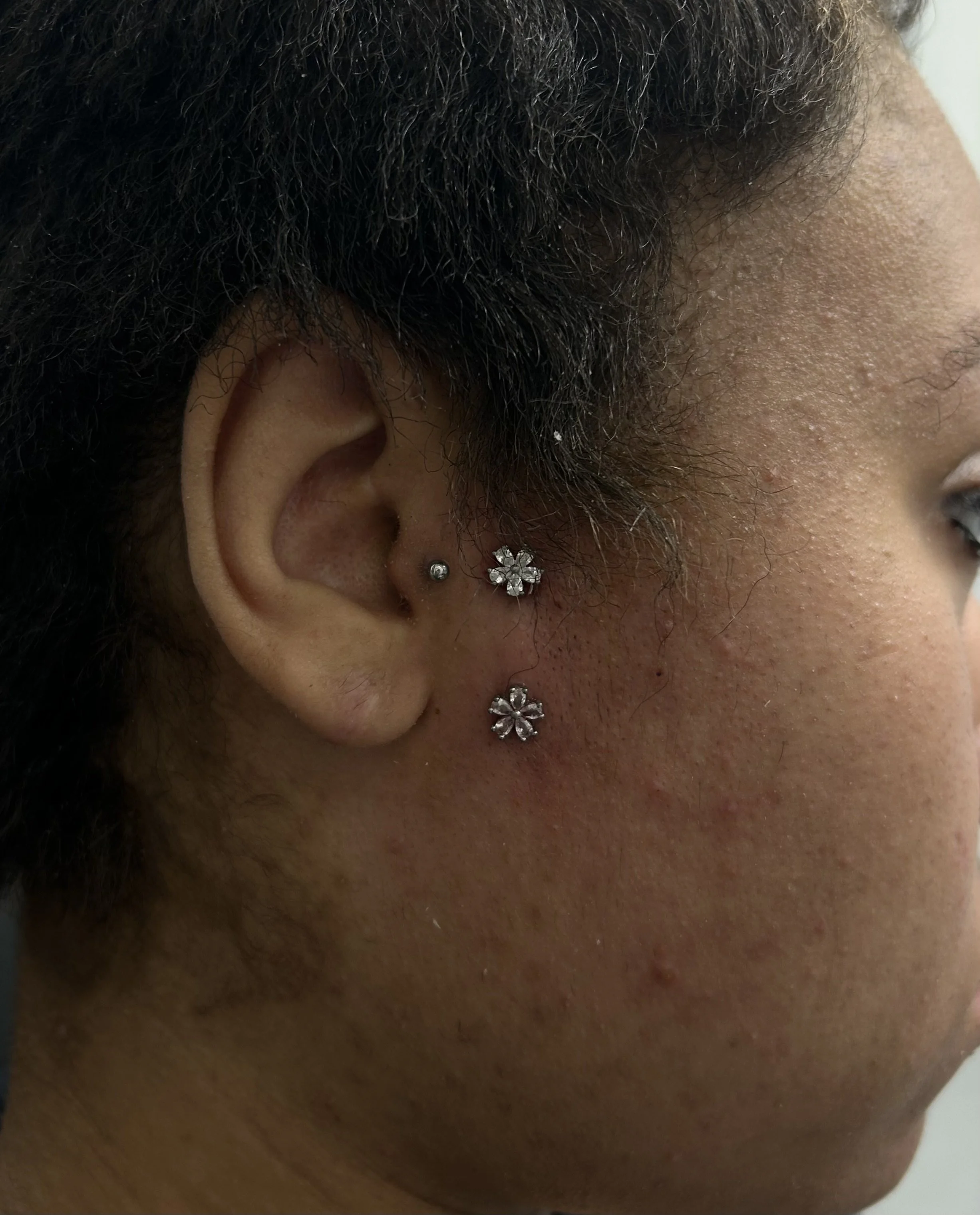 Close-up of a person's ear with multiple earrings, including a flower-shaped earring, a ball stud, and an ear cuff, with dark curly hair and a side of the face visible.